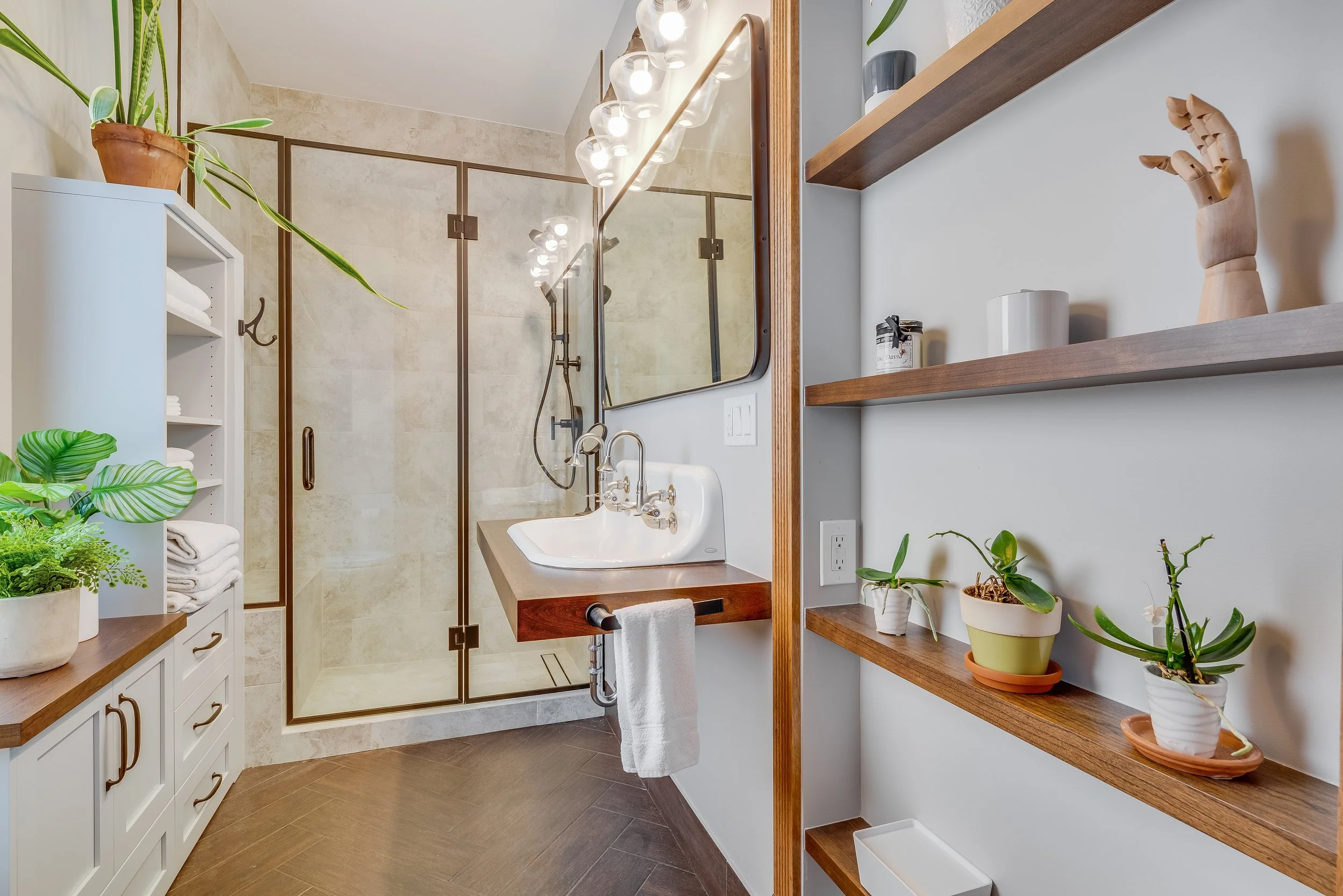 A modern bathroom with a glass-enclosed shower, a white sink with a wooden countertop, floating shelves with plants and decorative items, and a wooden cabinet with towels.