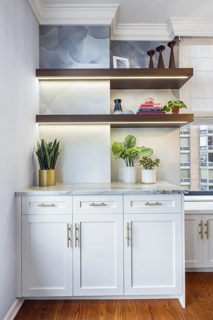 Interior of a modern kitchen with white cabinets, gold handles, a marble countertop, and two floating shelves with decorative items and plants. There is a large window on the right with a city view.