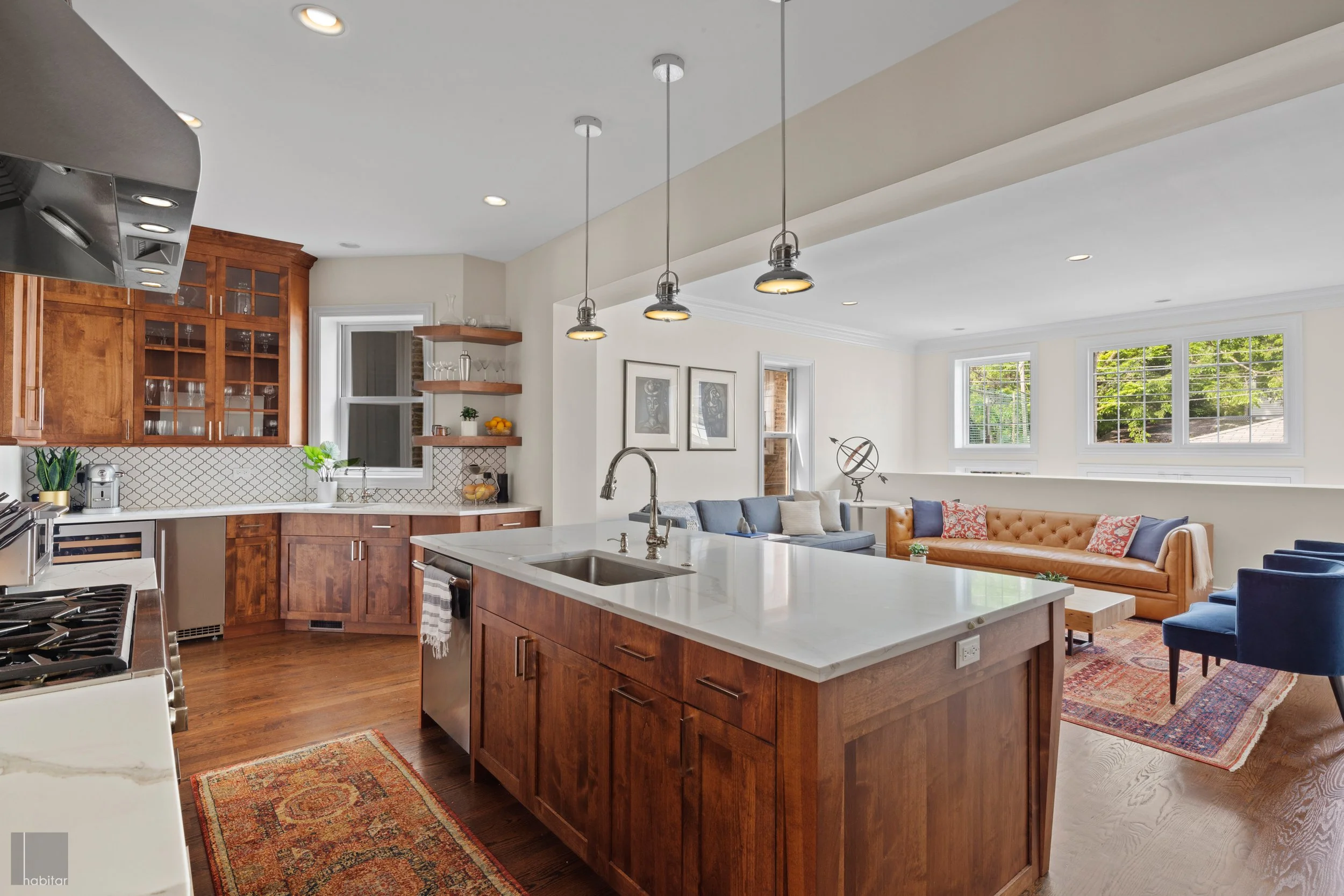 Open-concept kitchen and living room with wooden cabinets, white countertops, a gray sectional sofa, blue chairs, and large windows with a view of green trees.