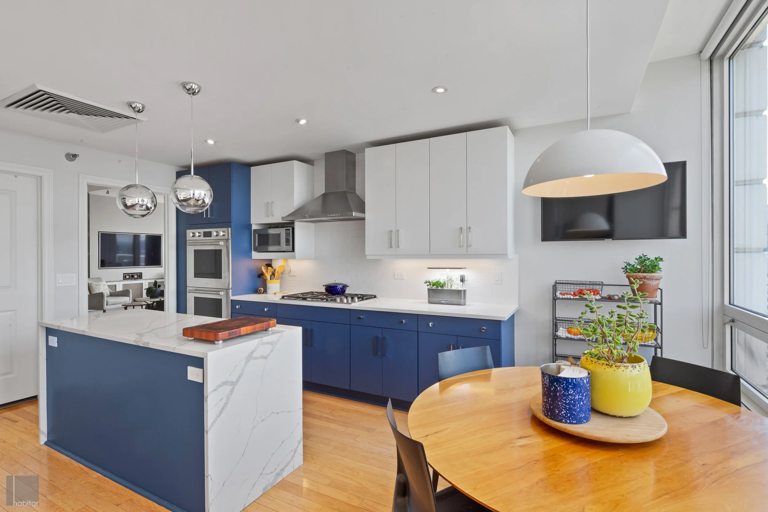 Modern kitchen with blue and white cabinets, a kitchen island with a marble pattern, a round wooden dining table with black chairs, potted plants, wall-mounted TV, and large windows bringing in natural light.