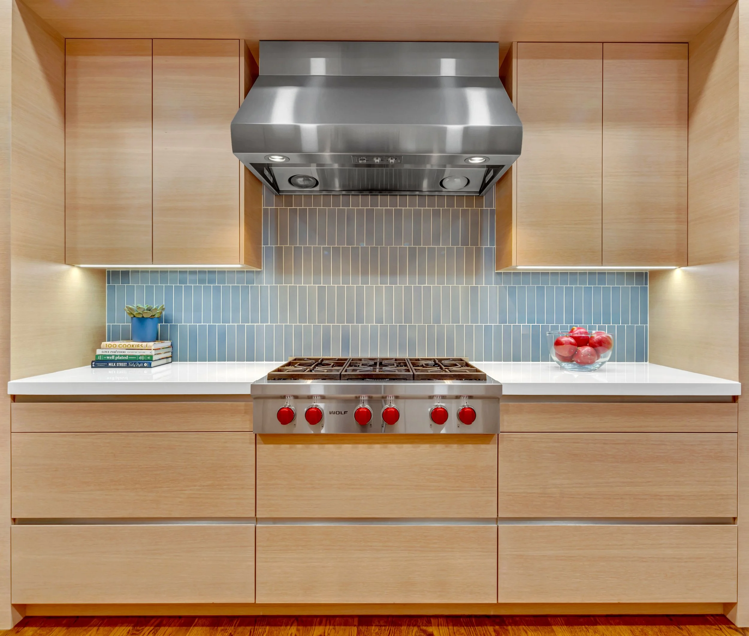 Modern kitchen with light wood cabinets, white countertop, blue tiled backsplash, stainless steel range hood, and stove with red knobs. Small potted plant and books on the left counter, bowl of red apples on the right.