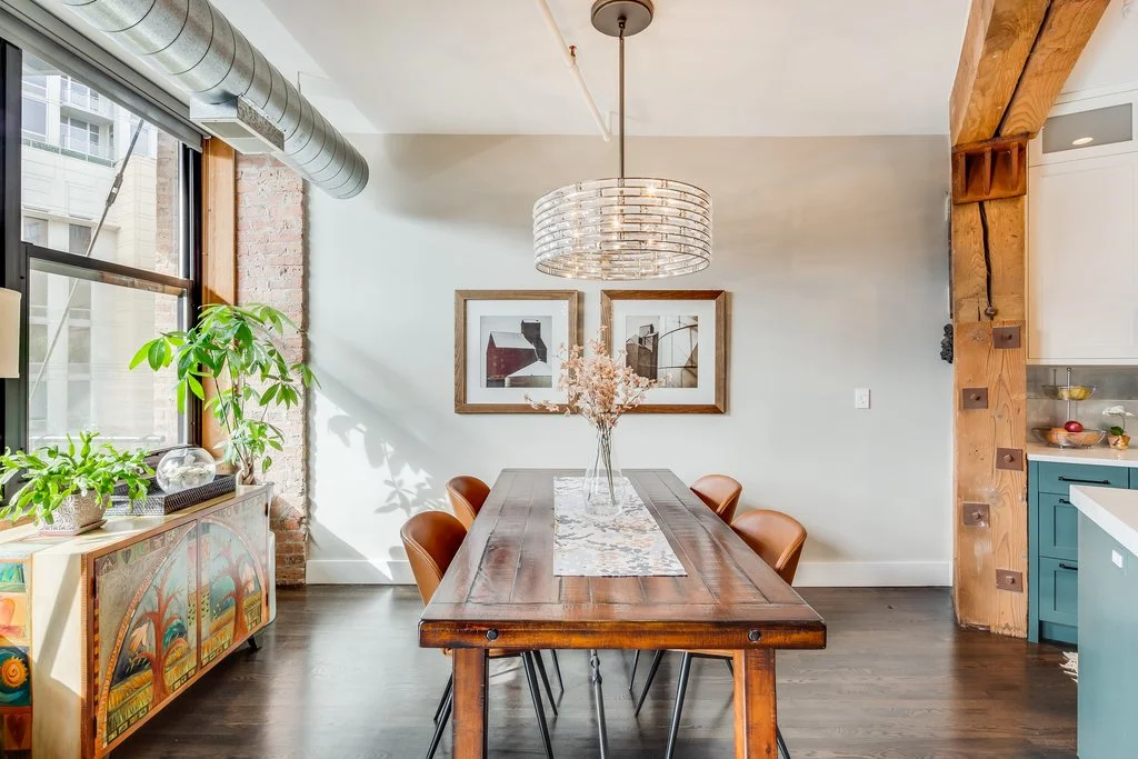 A modern dining room with a wooden table, six brown chairs, and a clear glass vase with dried flowers. Large window with a view of neighboring buildings, green plants on a decorated sideboard, two framed abstract artworks on the wall, and a contempor
