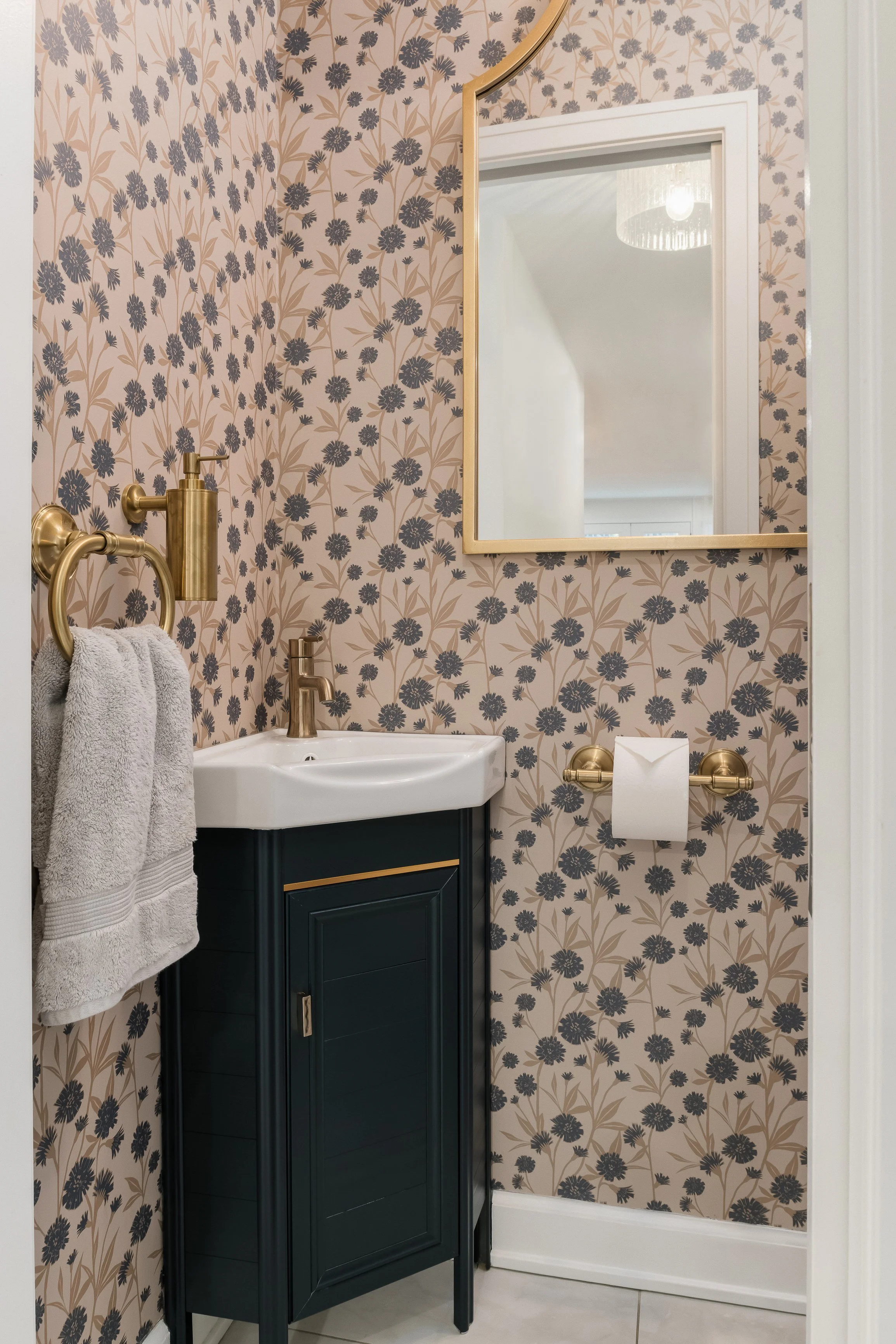 A small bathroom with floral patterned wallpaper, a mirror with a gold frame, a black vanity with a white sink, gold fixtures, a wall-mounted gold towel holder with a towel, and a toilet paper holder with a roll of toilet paper.