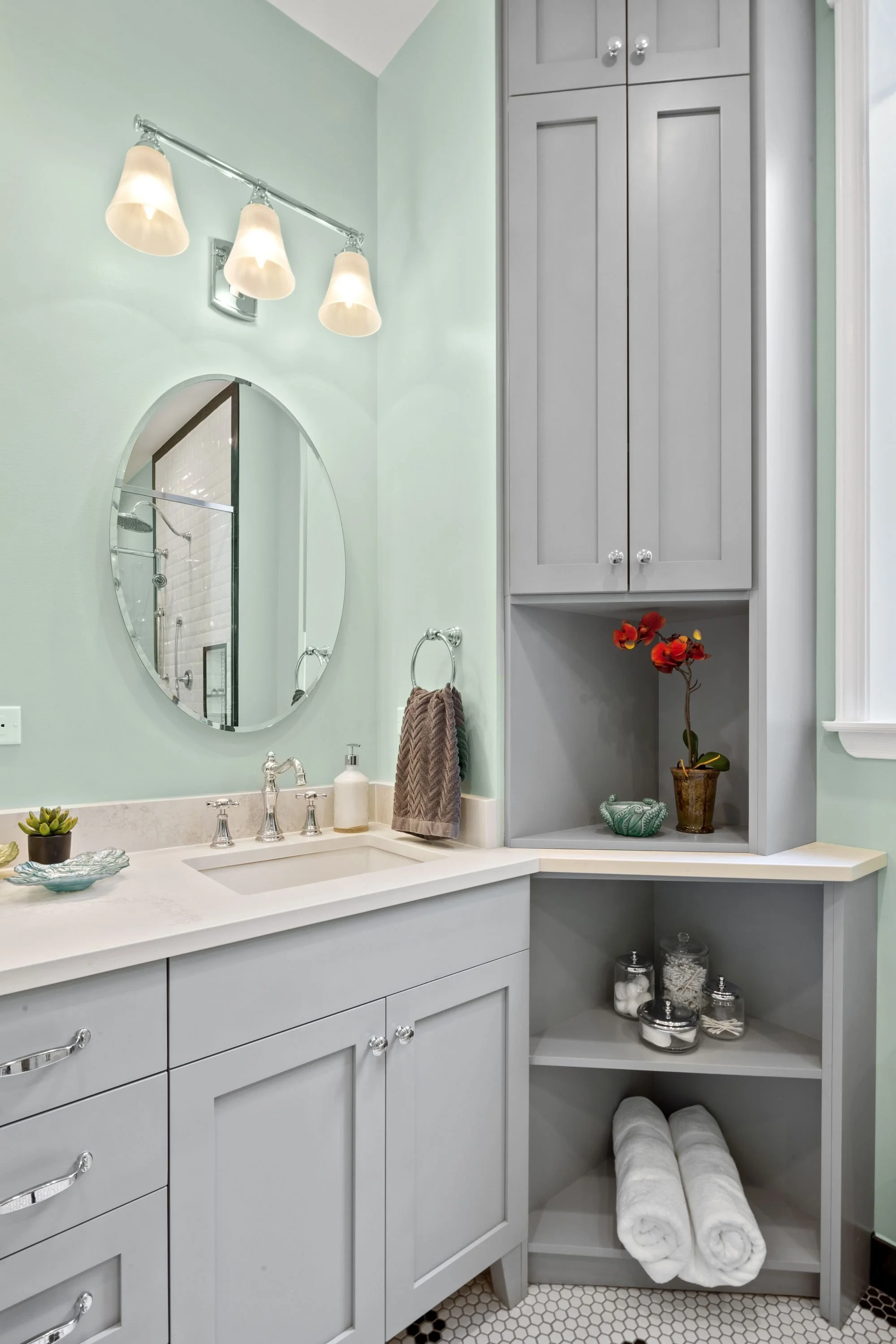 Bathroom with light green walls, white countertop vanity, round mirror, three-paneled storage cabinet, flowers in a pot, rolled white towels, jars with bathroom items, and a hexagonal tile floor.