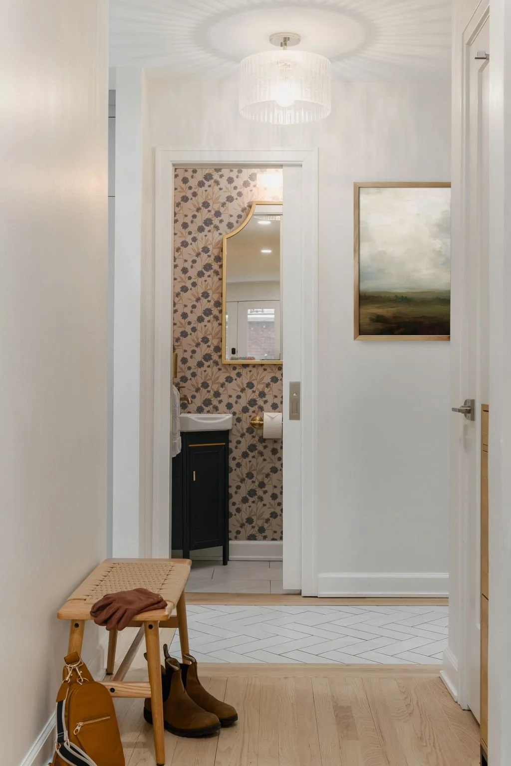 View of an entryway leading to a small bathroom with floral wallpaper, a mirror, and a white sink. The hallway has white walls, a modern light fixture, and a wooden bench with a pair of brown boots and a mustard-colored bag placed nearby.