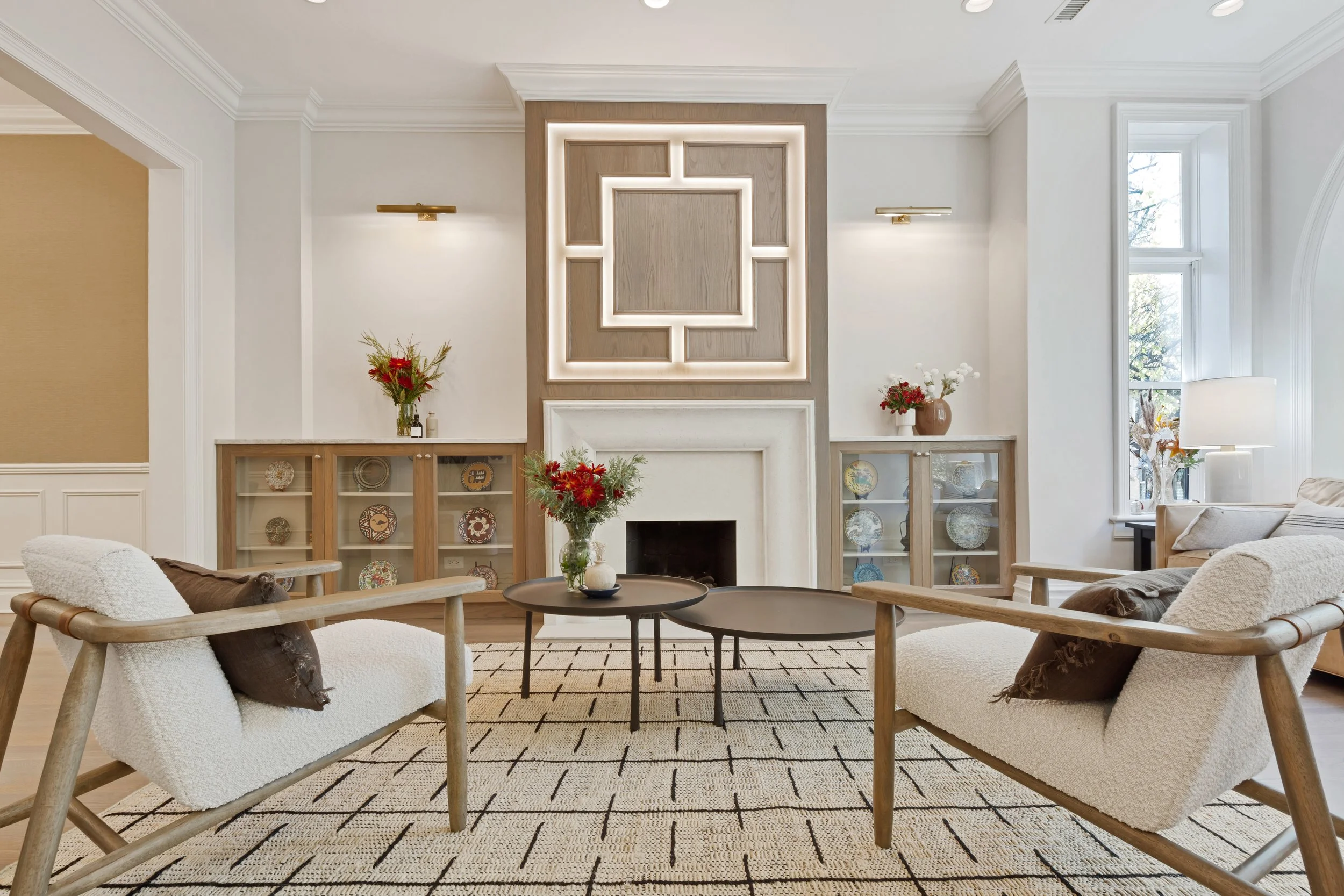 Modern living room with two cream-colored armchairs, a black coffee table with vases of flowers, a fireplace with decorative wood paneling and backlighting, wooden display cabinets with plates, and large windows with white curtains.
