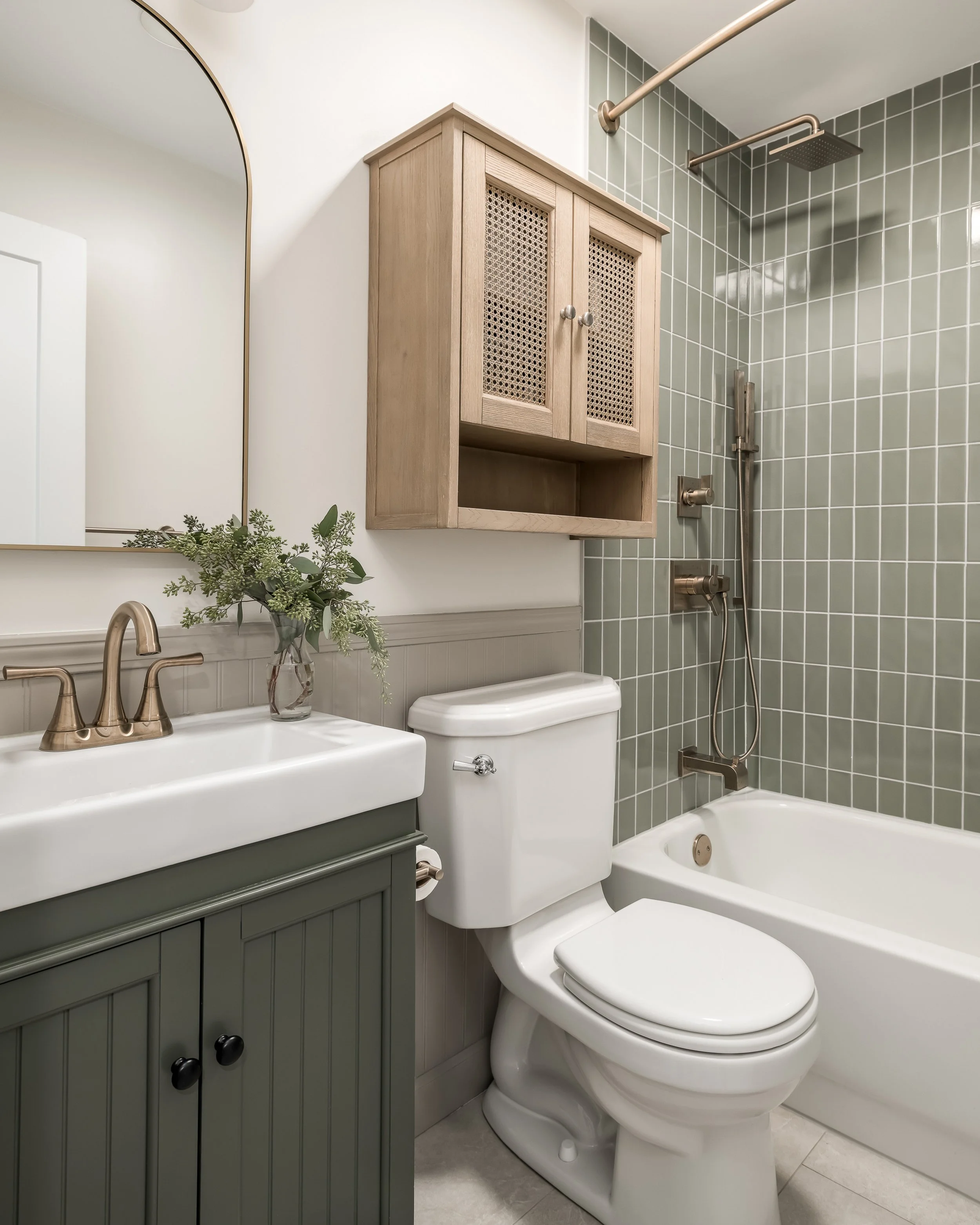 Bathroom with green tiled shower area, white bathtub, white toilet, gray vanity with a white sink, a mirror, a small woven wall cabinet, and a vase with green foliage on the vanity.