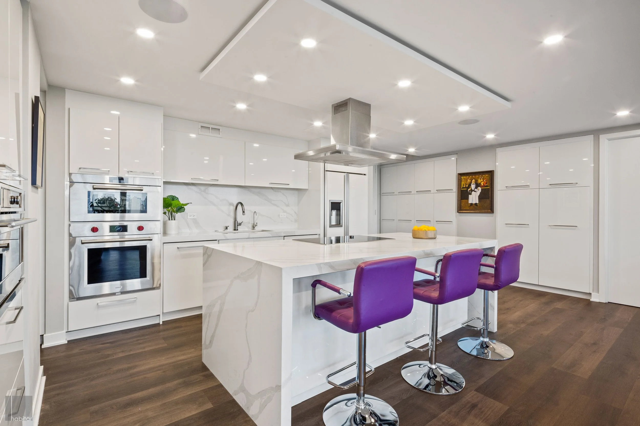 Modern kitchen with white cabinets and marble backsplash, large marble island with purple bar stools, built-in oven, and stainless steel range hood.