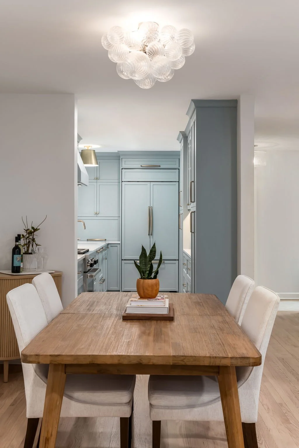 A dining room with a wooden table and white chairs. On the table, there is a potted plant and a stack of books. The kitchen in the background has light blue cabinetry and modern appliances. A decorative chandelier is hanging from the ceiling.