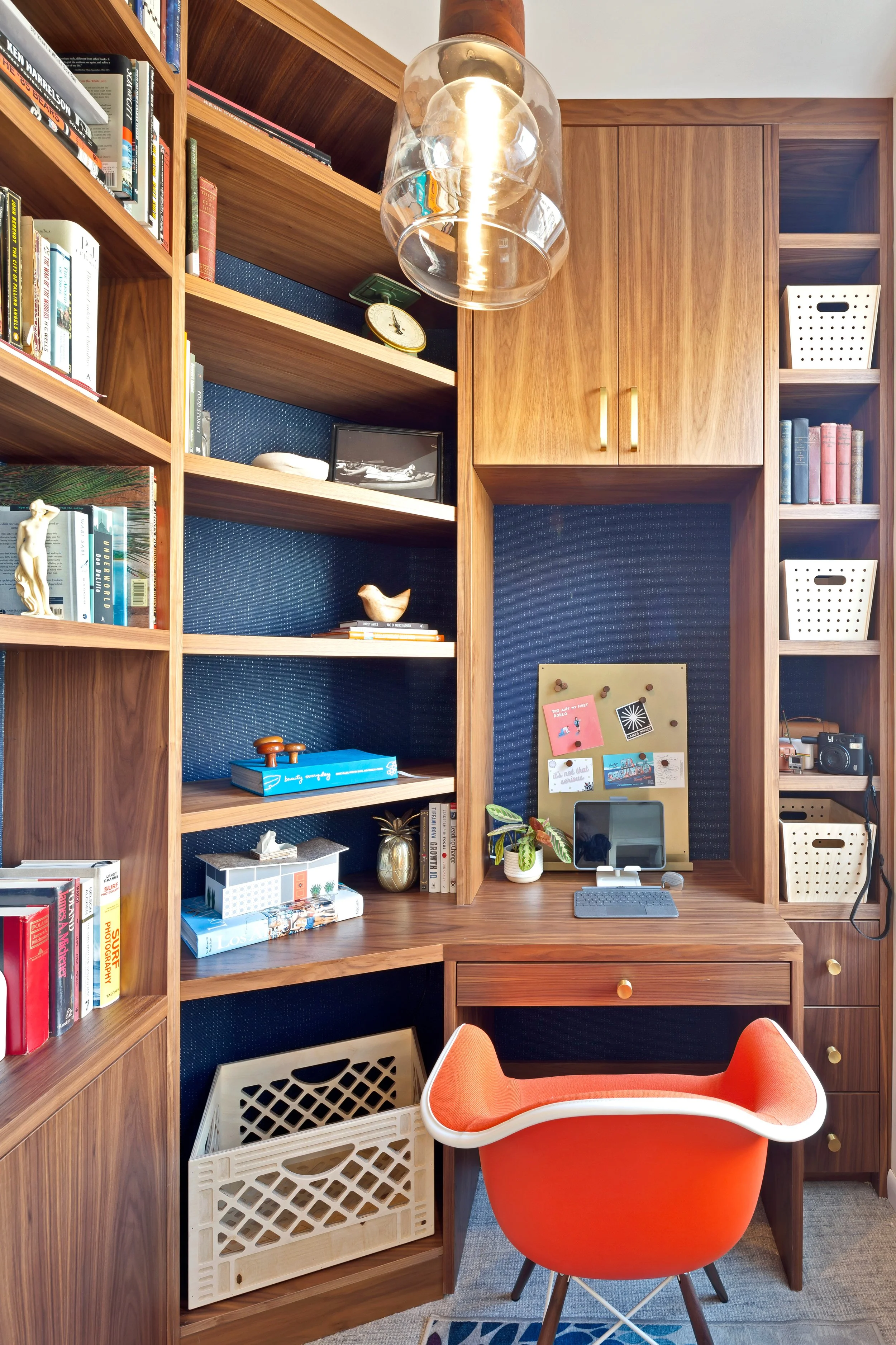 Home office with built-in wooden shelves, a small desk with a monitor and plants, and a modern orange chair in front of the desk.