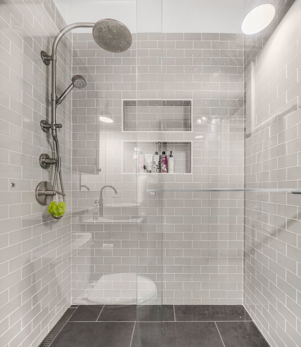 Enclosed shower with white subway tile, a large overhead rain showerhead, and built-in shelves with toiletries.
