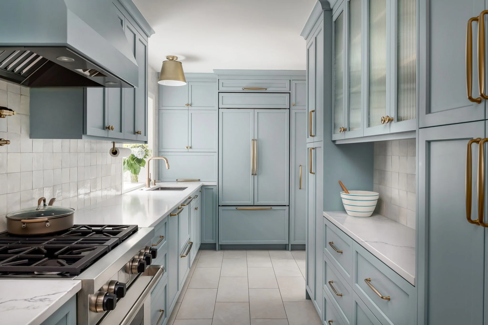 Light blue kitchen with brass hardware, white countertops, stove, built-in refrigerator, cabinets, a window with a plant, and a white tiled backsplash.