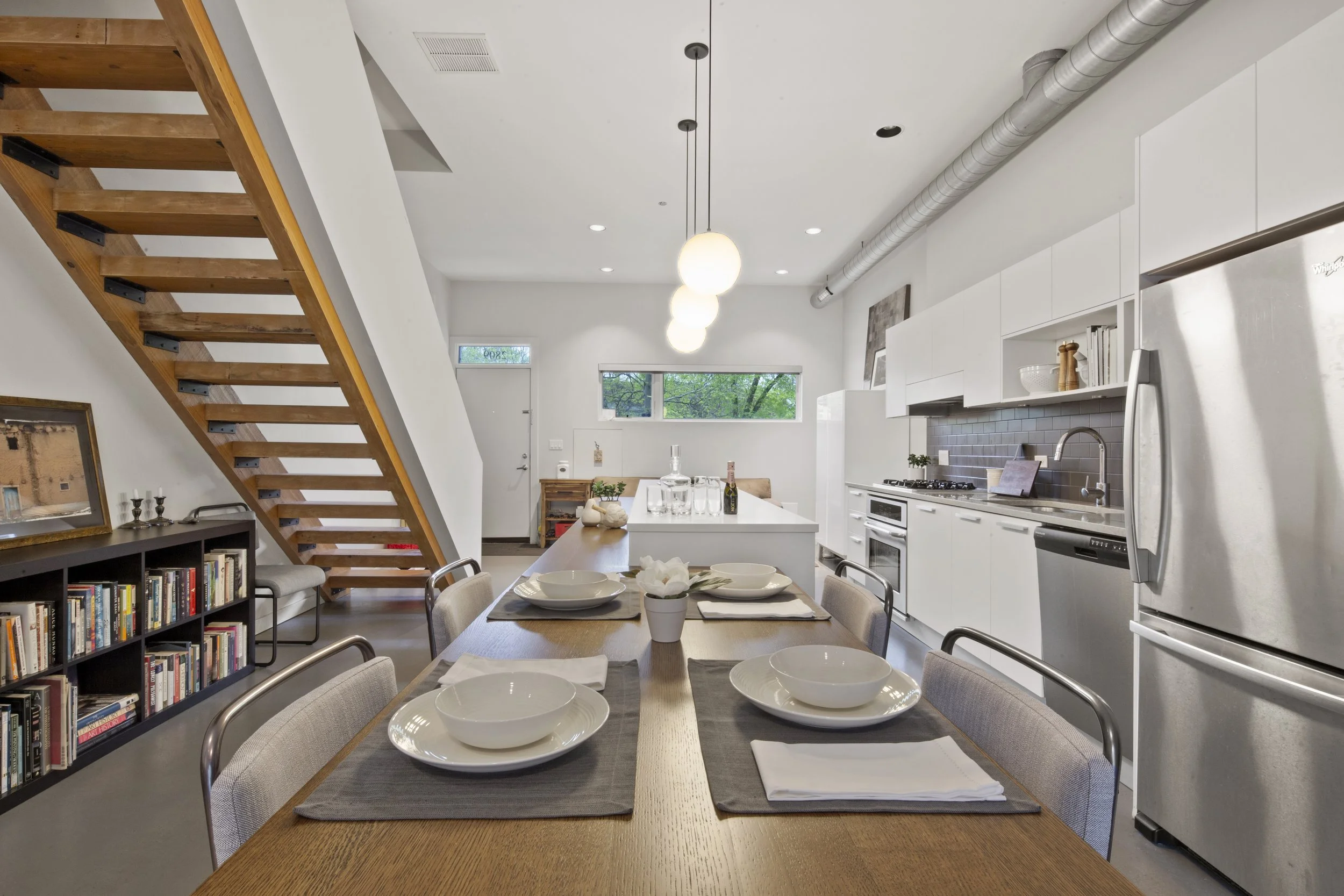 Modern open-concept kitchen and dining area with a wooden table set with white dishes, a cozy white kitchen island, stainless steel appliances, and a staircase leading upstairs.