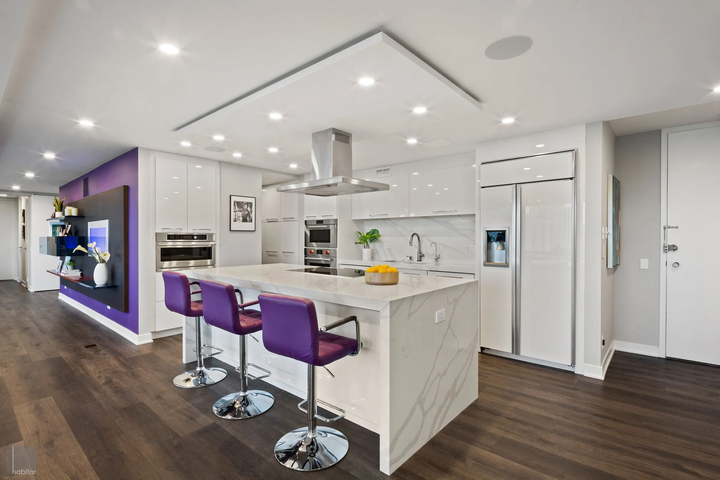 Modern kitchen with white cabinets, marble island, purple bar stools, stainless steel appliances, and wooden flooring.