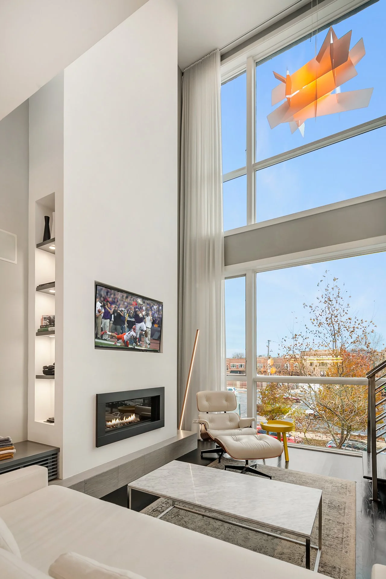 Modern living room with large windows, white walls, a mounted TV showing a football game, a white armchair with a matching ottoman, a small yellow side table, a marble-top coffee table, curtains, and a ceiling lamp outside the window.