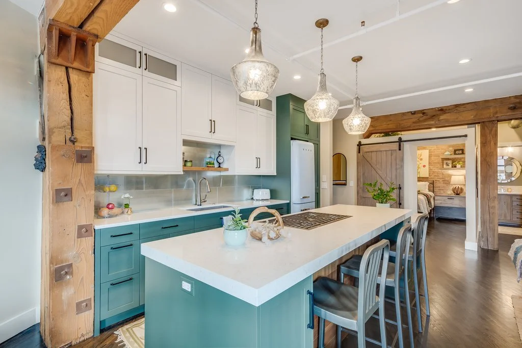 Modern kitchen with a large island, white and teal cabinetry, wooden accents, and pendant lighting, with a view into a bedroom area.