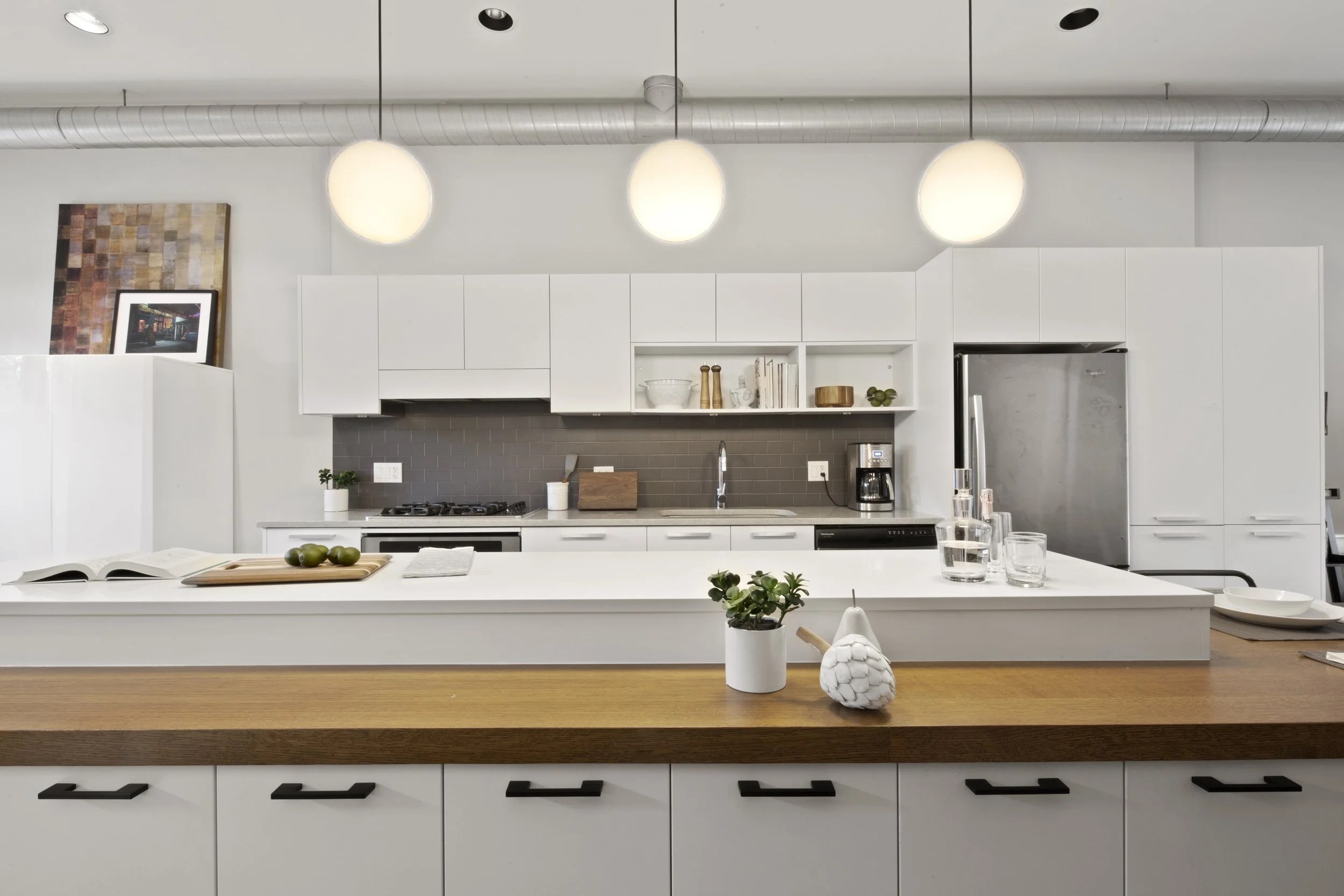 Modern white kitchen with wooden countertop island, gray backsplash, ceiling lights, and stainless steel appliances.