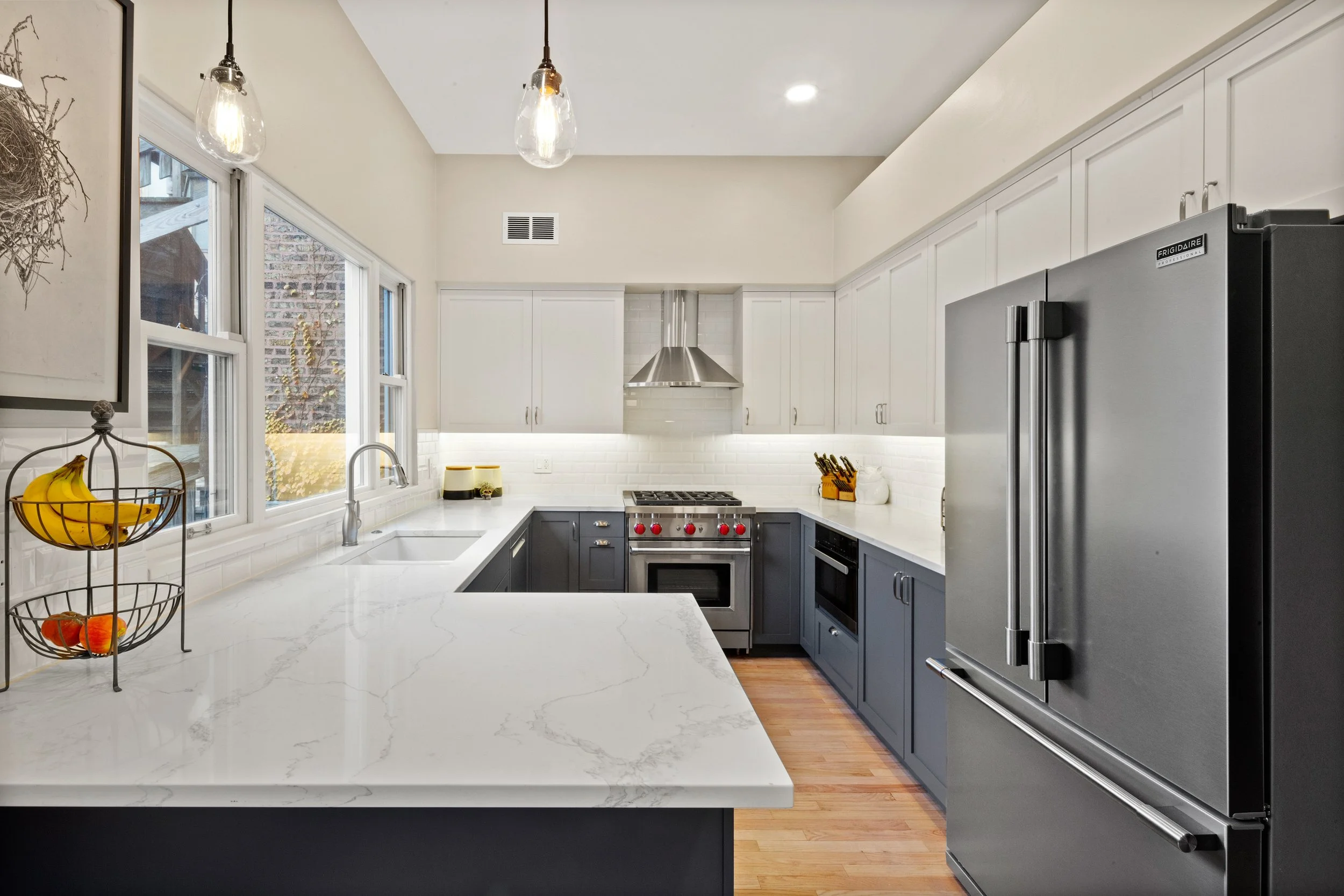 Modern kitchen with white upper cabinets, dark lower cabinets, a stainless steel refrigerator, a white marble countertop, a gas stove, and pendant lighting.