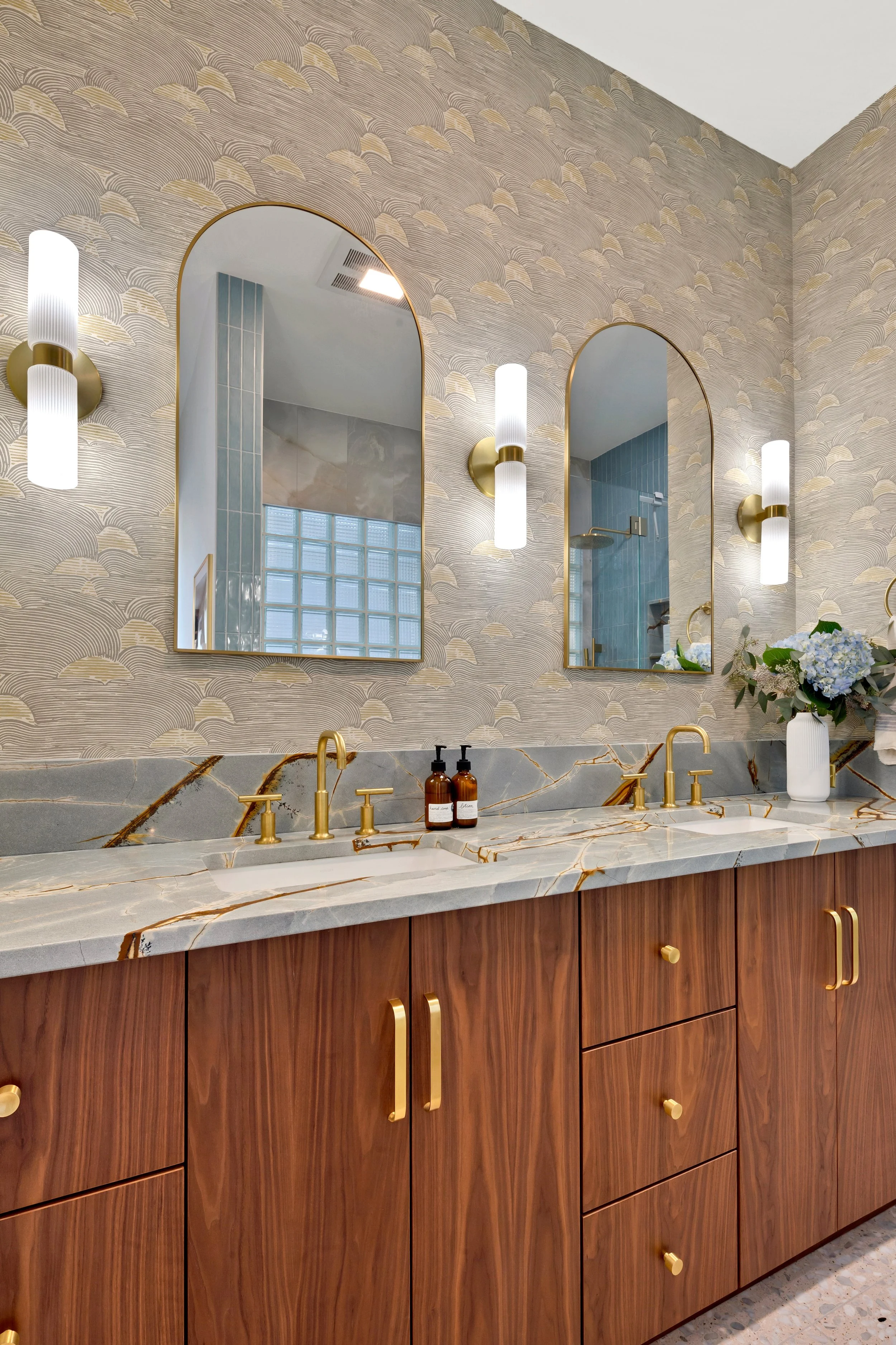 A modern bathroom with a marble countertop, gold fixtures, and two arched mirrors above the sinks. The wall has a patterned wallpaper, and there are wall-mounted lights on either side of each mirror. A vase with blue and white flowers is on the right