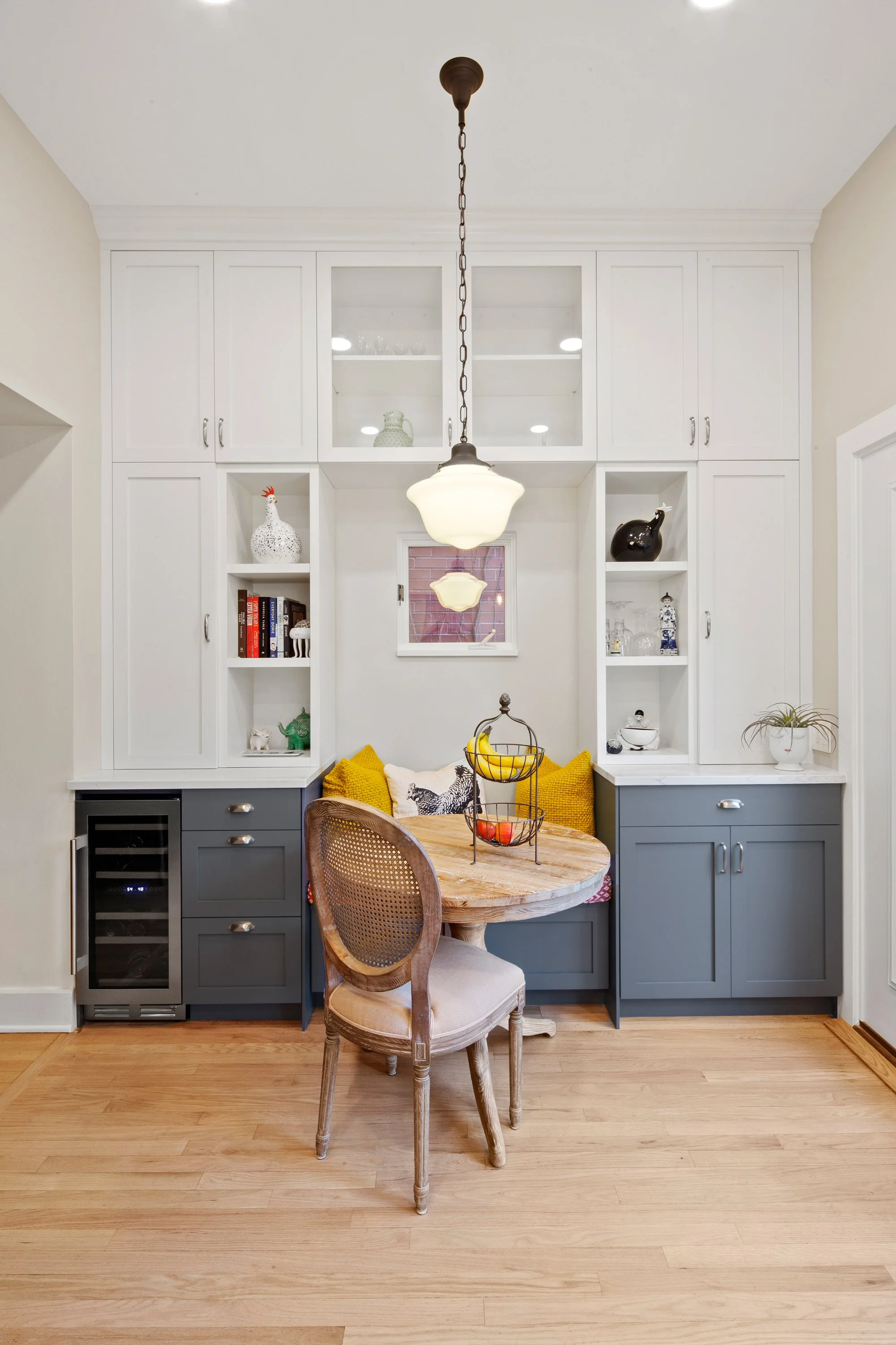 A kitchen nook with a round wooden table, vintage wood chair, built-in white and gray cabinets, and decorative items including a fruit basket with bananas and apples, and shelves with books and ceramics.