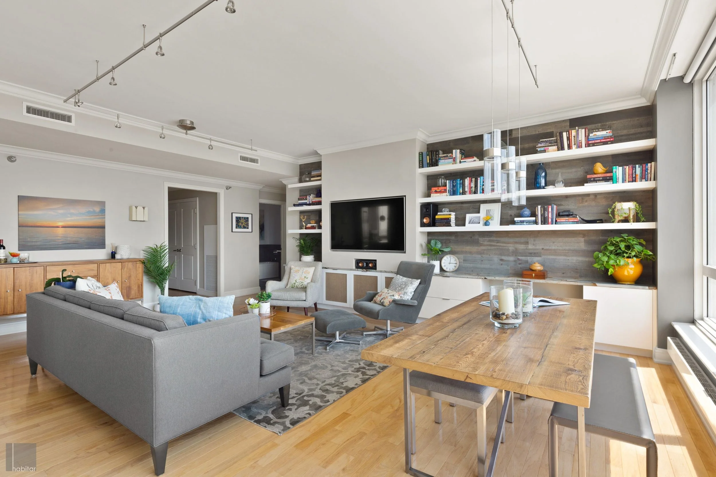 A modern living room with a gray sofa, two gray chairs, a wooden coffee table, a large built-in bookshelf with various books and decorative items, a wall-mounted TV, and a wooden dining table with gray chairs. Large windows let in natural light, and 