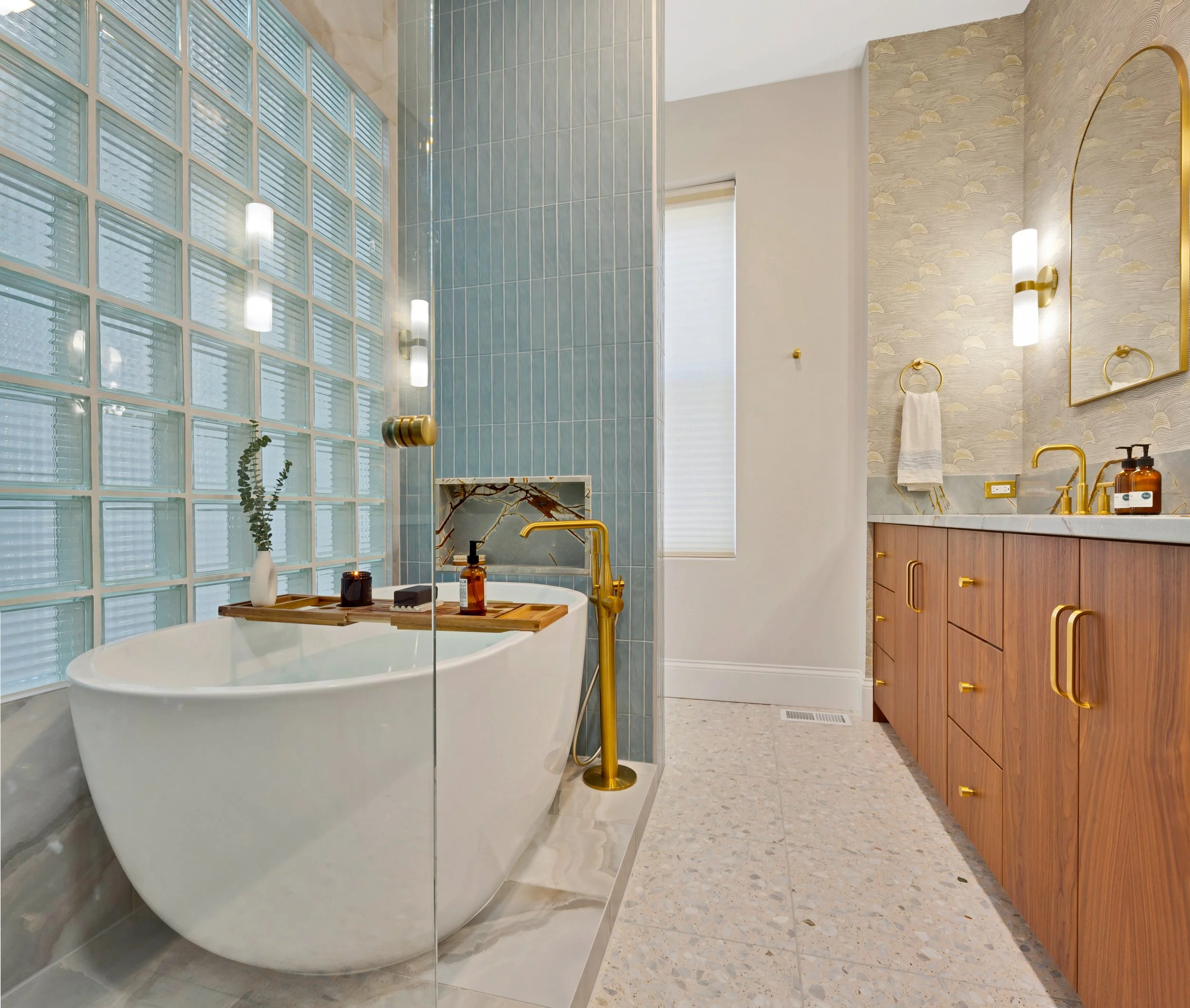 Modern bathroom with a freestanding bathtub, a glass wall, blue tiles, wood vanity with gold fixtures, and decorative lighting.