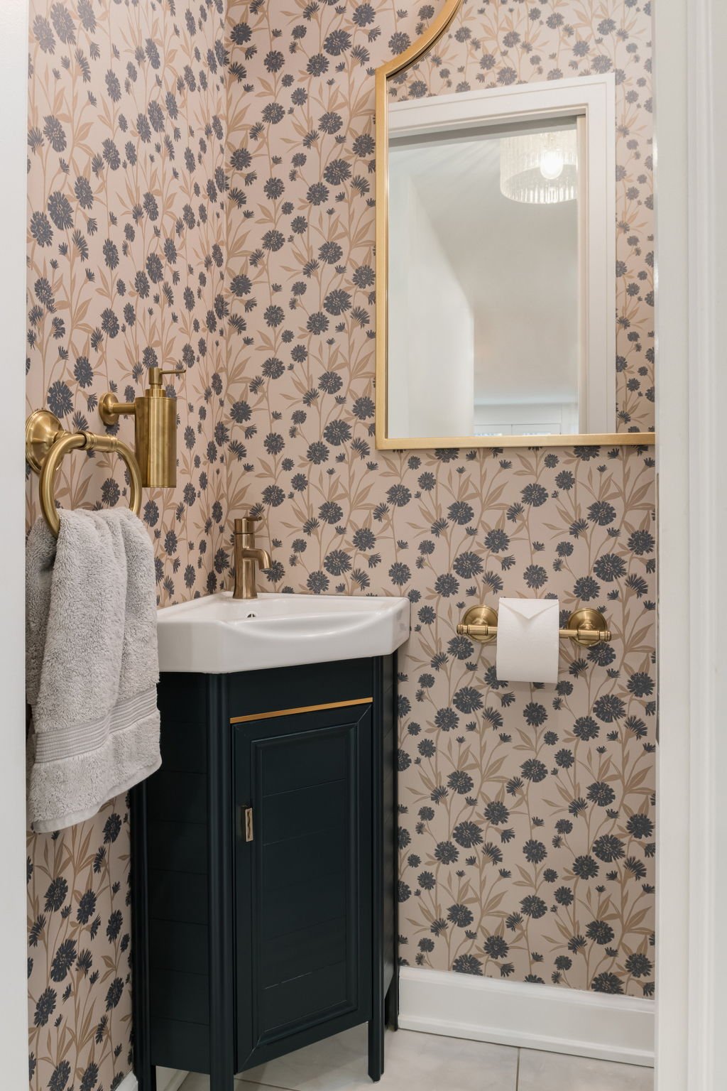 Bathroom with floral patterned wallpaper, a small black vanity with gold accents, a white sink, a gold-mounted towel ring with a gray towel, a gold wall-mounted toilet paper holder, a mirror with a gold frame, and a partially visible light fixture.