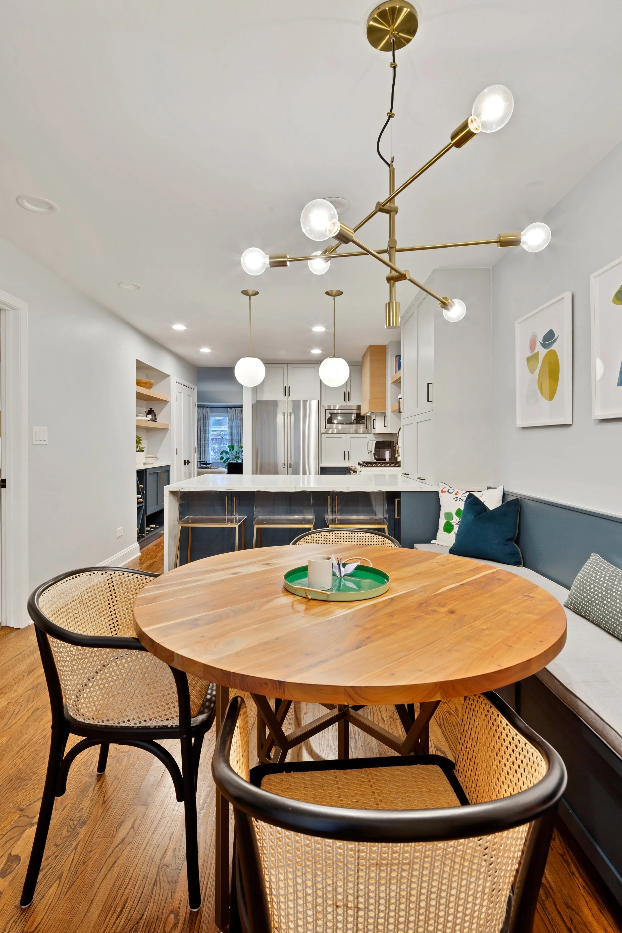 Modern kitchen and dining area with a wooden table surrounded by wicker chairs, a navy blue bench with pillows, a gold chandelier with exposed bulbs, and a white ceiling with recessed lighting.