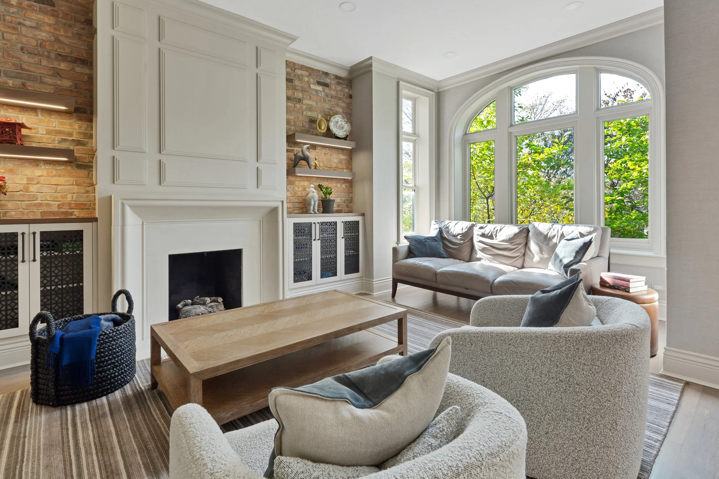 Living room with large bay windows, white sofa, armchair, wooden coffee table, and brick fireplace with built-in shelves and cabinetry.
