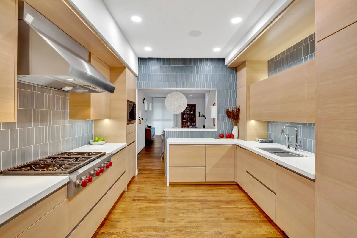 Modern kitchen with light wood cabinets, white countertops, and gray tiled backsplash. The kitchen has a gas stove on the left and a double sink on the right, with a view into a living area with wooden floors and a white spherical pendant light.