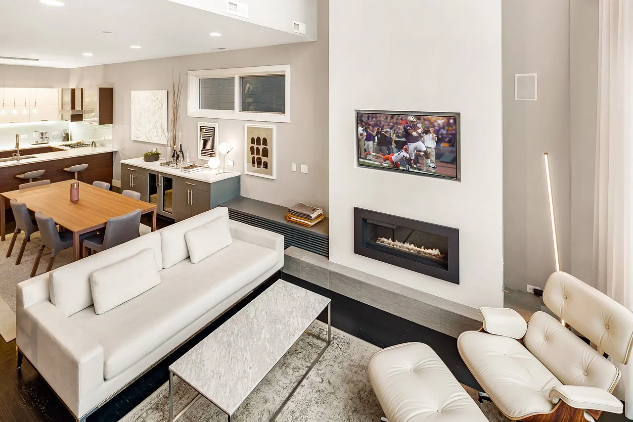 Modern living room with white couch, white armchair, wall-mounted TV showing a football game, and a fireplace beneath the TV. Dining area with wooden table and gray chairs, kitchen in the background.