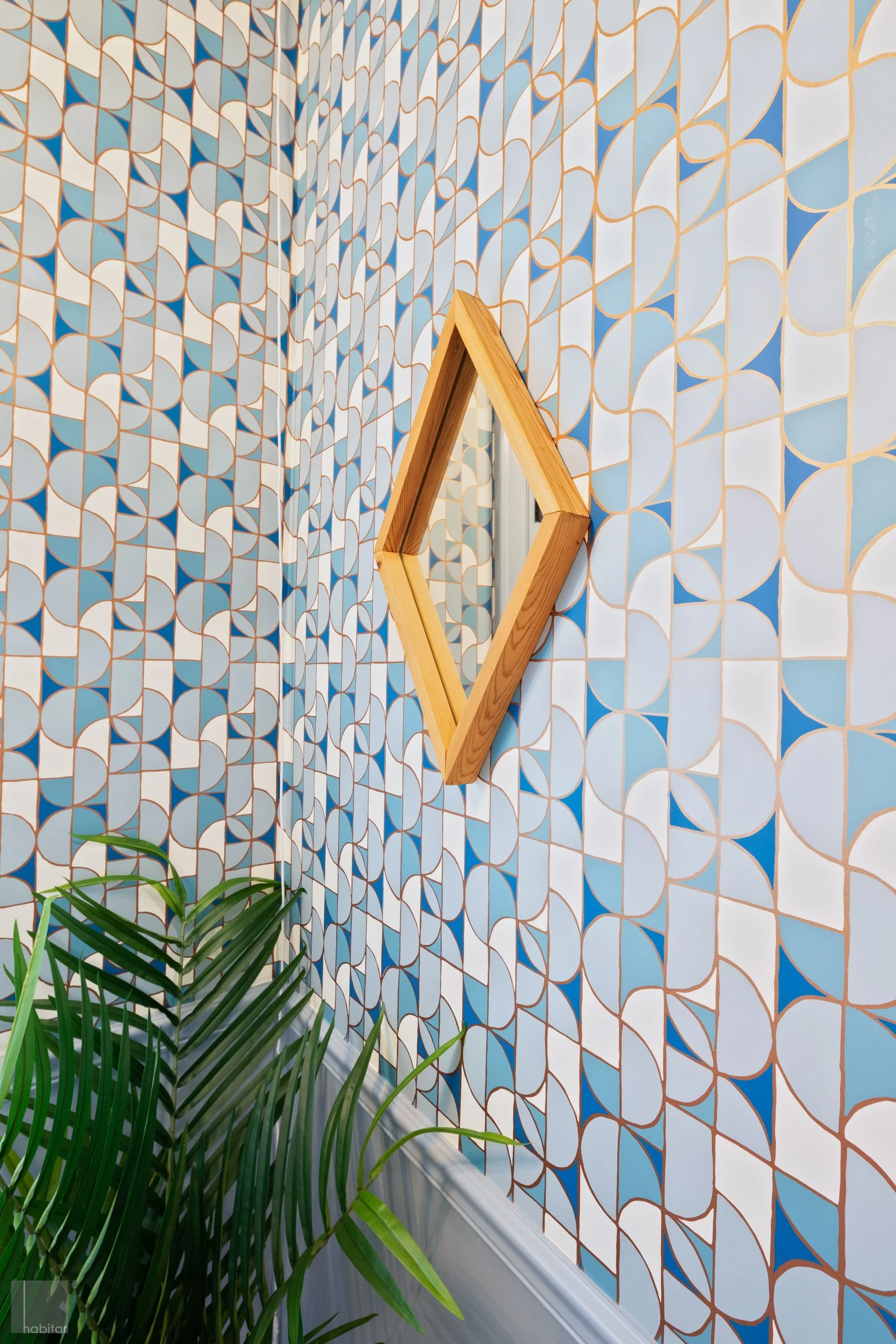 A decorative wall with geometric blue, white, and gold patterned tiles, a wooden-framed mirror in a diamond shape, and a green leafy plant in the bottom left corner.