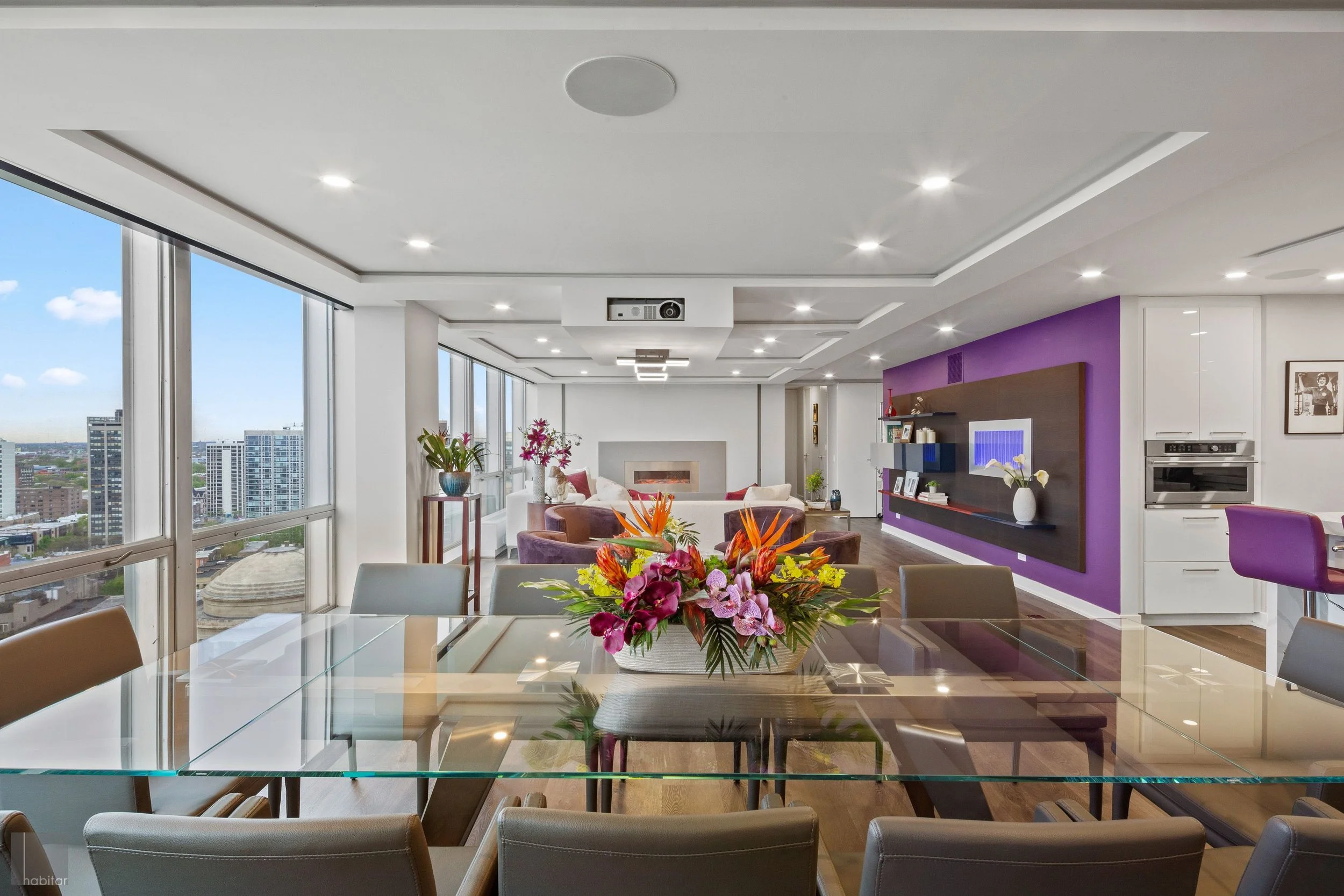 Modern high-rise apartment living room with large windows, colorful furniture, a flower centerpiece on a glass dining table, and a view of city buildings outside.