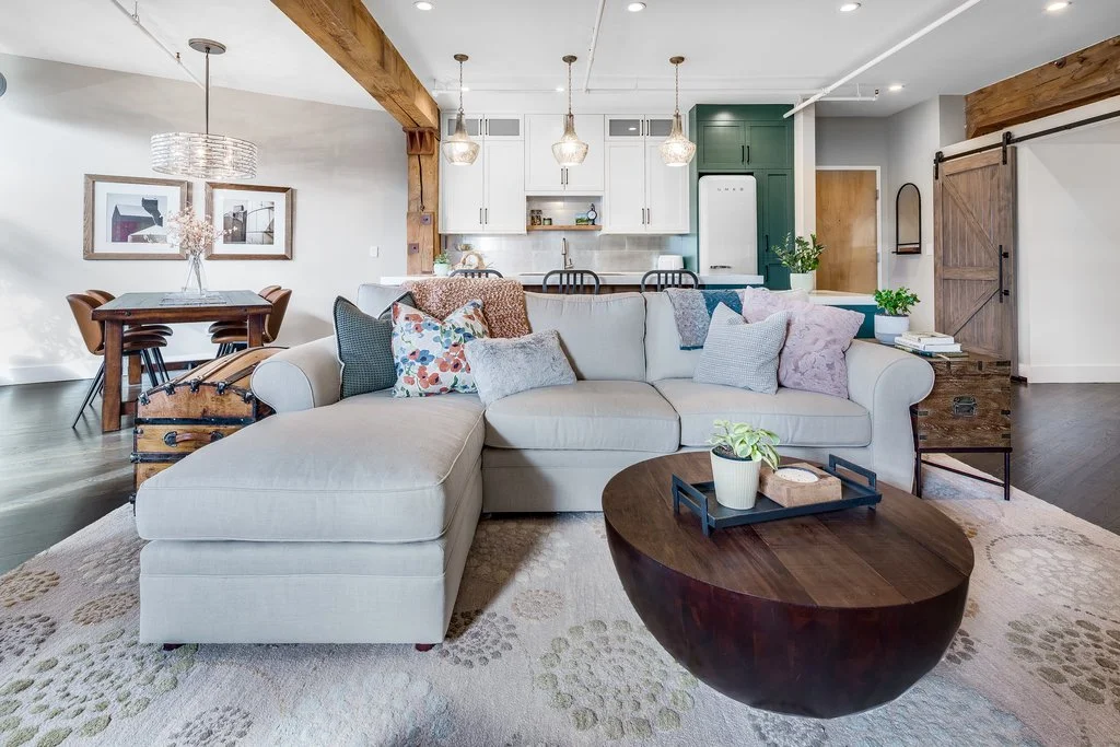 Open-concept living room and kitchen with a beige sectional sofa, colorful throw pillows, wooden coffee table, dining area with a wooden table and six chairs, white cabinets, green accent wall, pendant lights, and farmhouse-style barn doors.
