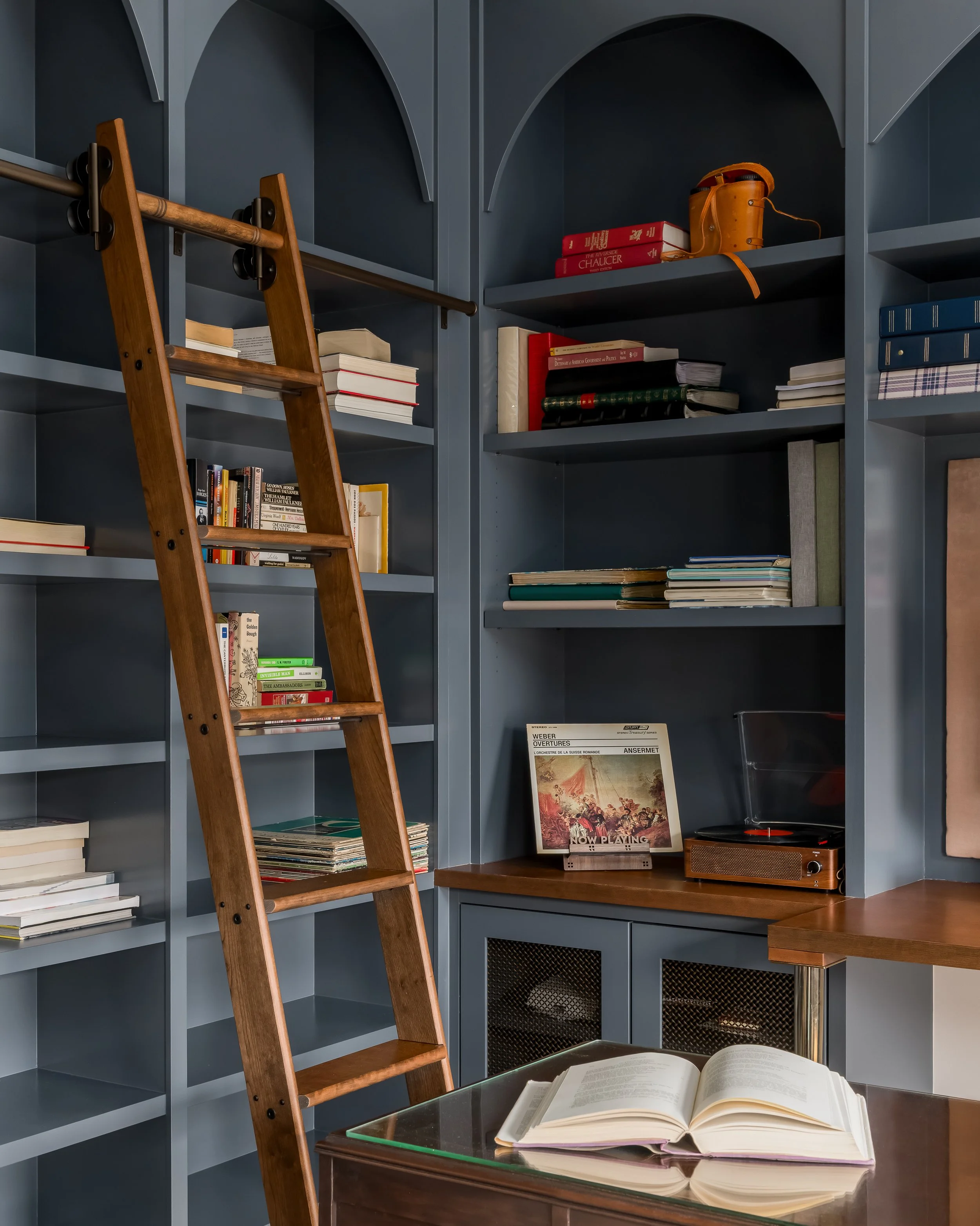 Interior of a room with blue bookshelves filled with books, a ladder leaning against the shelves, a record player on a wooden cabinet, and an open book on a glass table.