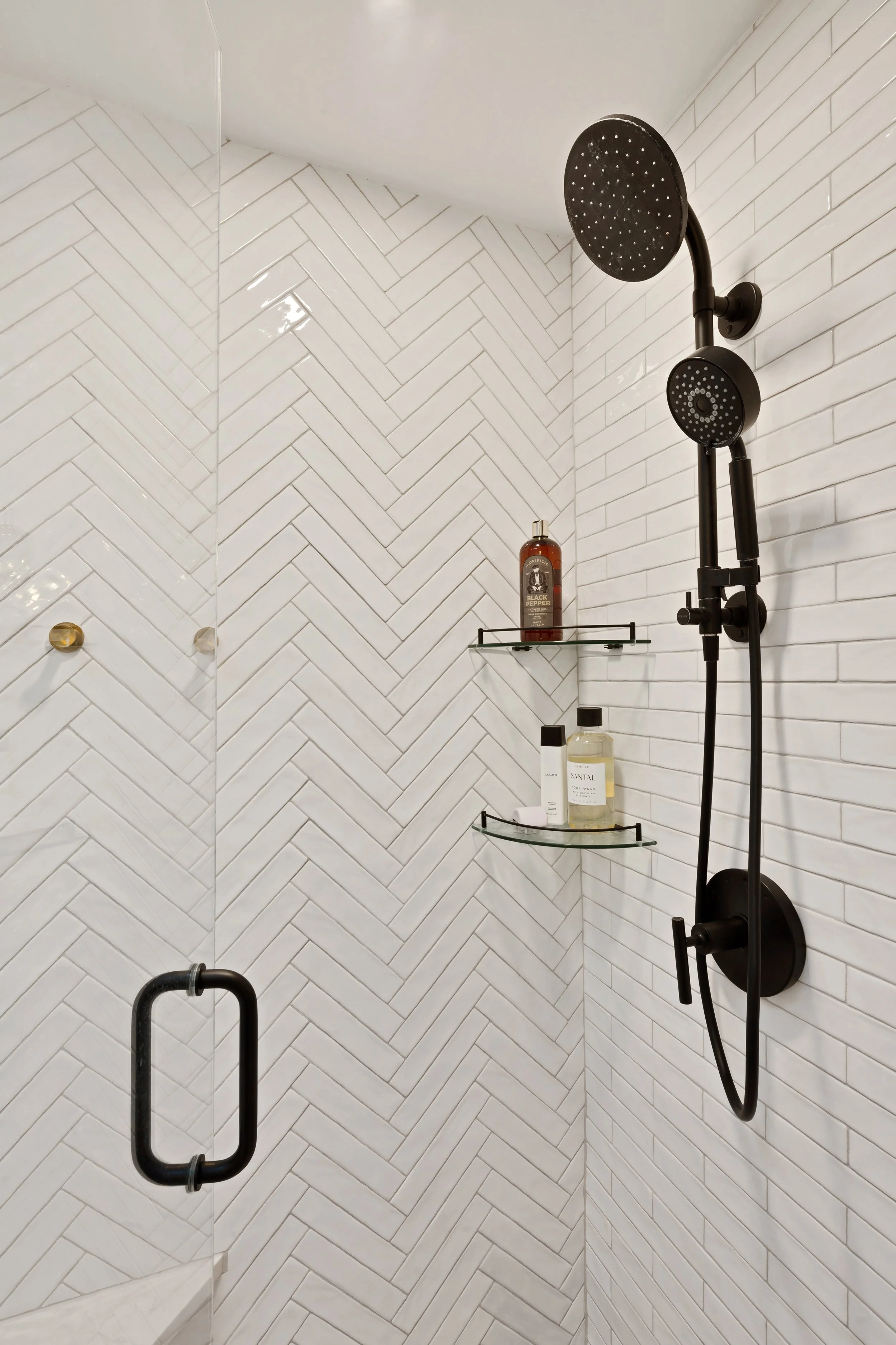 White herringbone tile shower with black showerhead and control, glass shelf with bottles of shampoo and body wash.