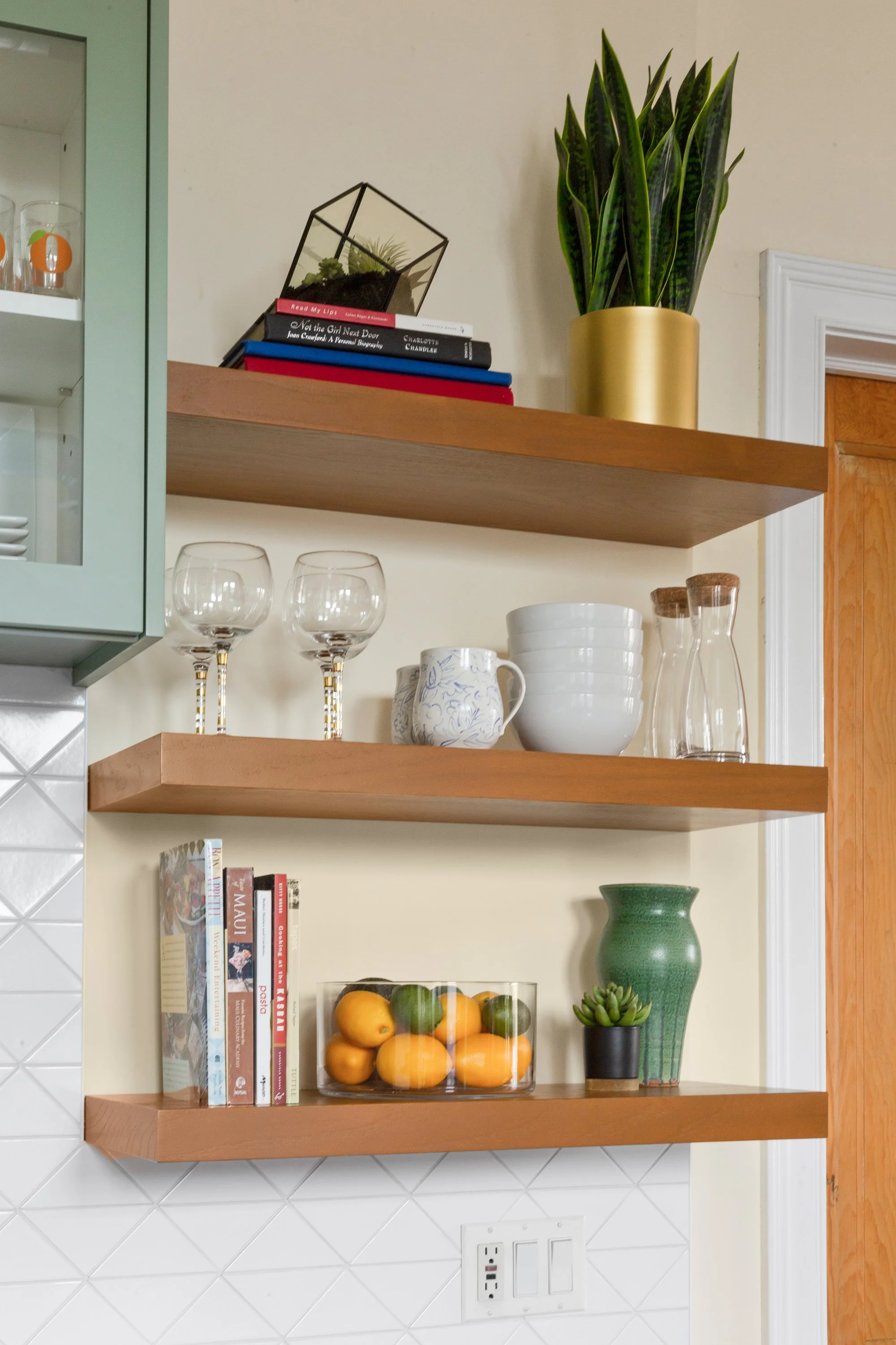 Wooden shelves with books, glassware, bowls, a pitcher, a vase, and a small potted plant on a white tiled wall.