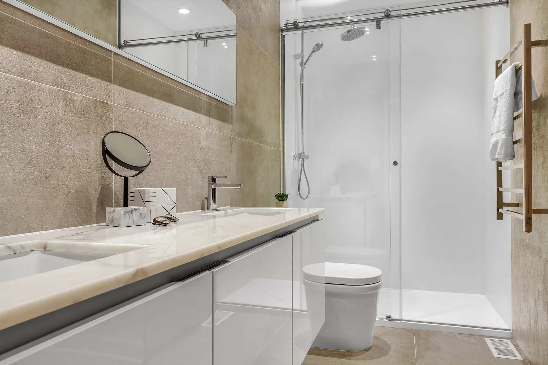 Modern bathroom with beige tiled wall, white countertop with sink, small mirror, and decorative plant. Shower with glass door and rainfall showerhead, wooden towel rack with towel, white toilet, and ventilation grill on beige floor.