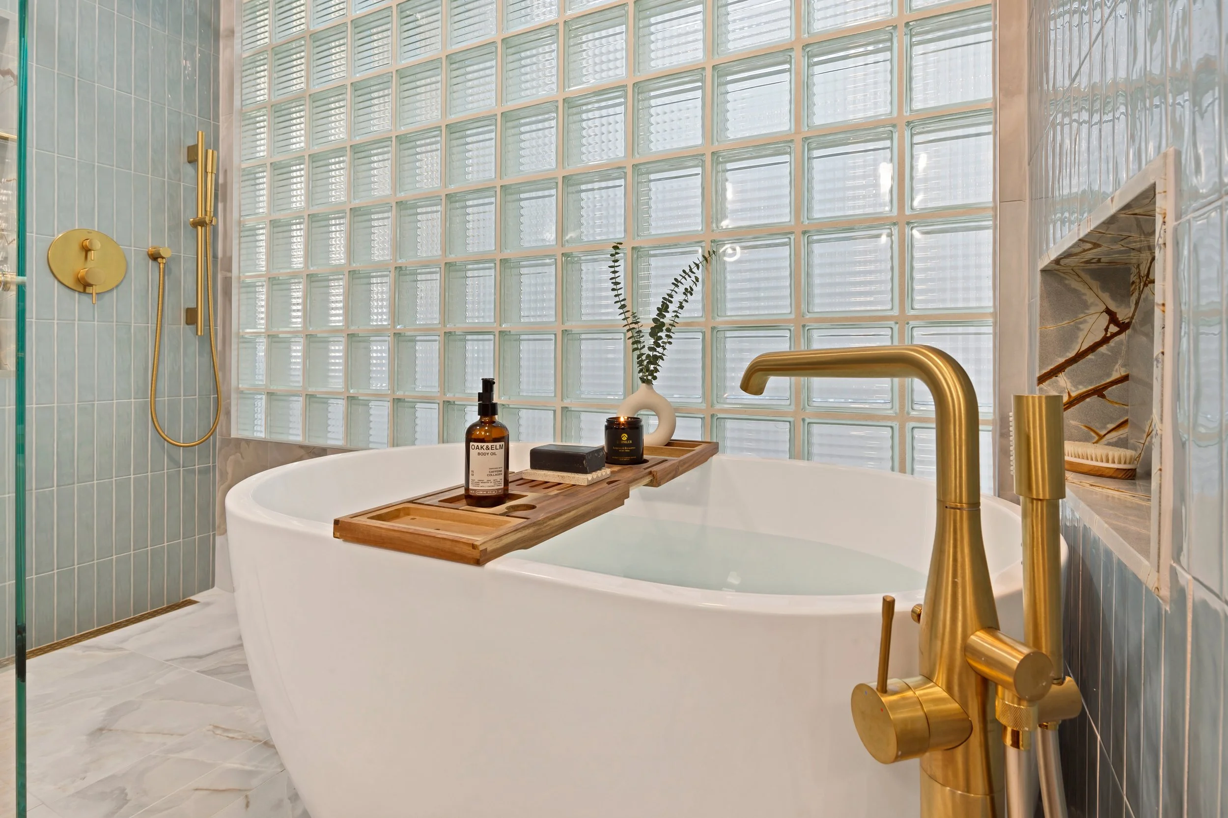 Modern bathroom with a white bathtub, gold fixtures, and glass block wall. A wooden bath tray holds bottles, a candle, and a decorative vase with greenery.