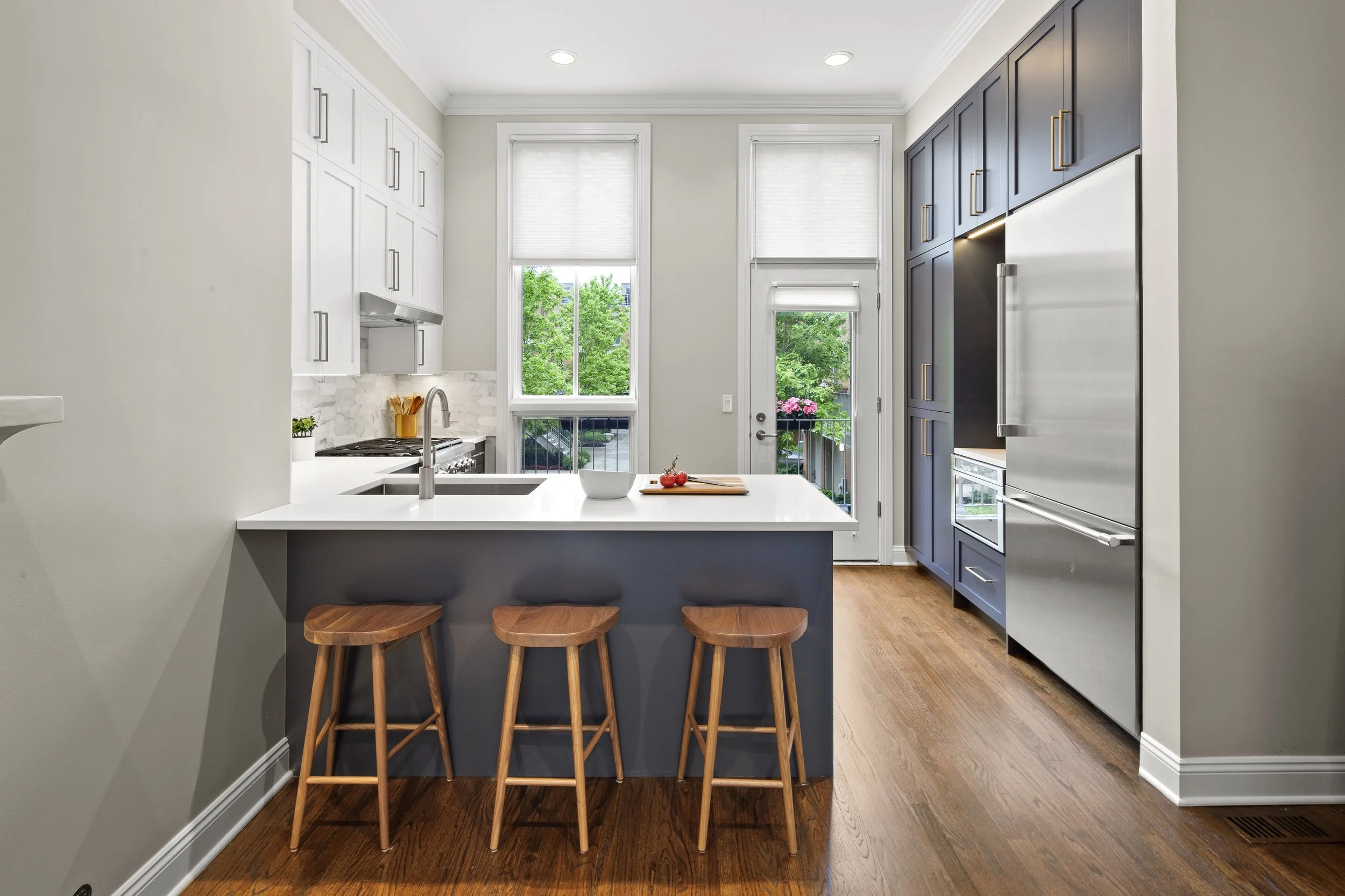 Modern kitchen with white countertops, dark blue cabinets, wooden stools, stainless steel appliances, and large windows with a view of green trees outside.