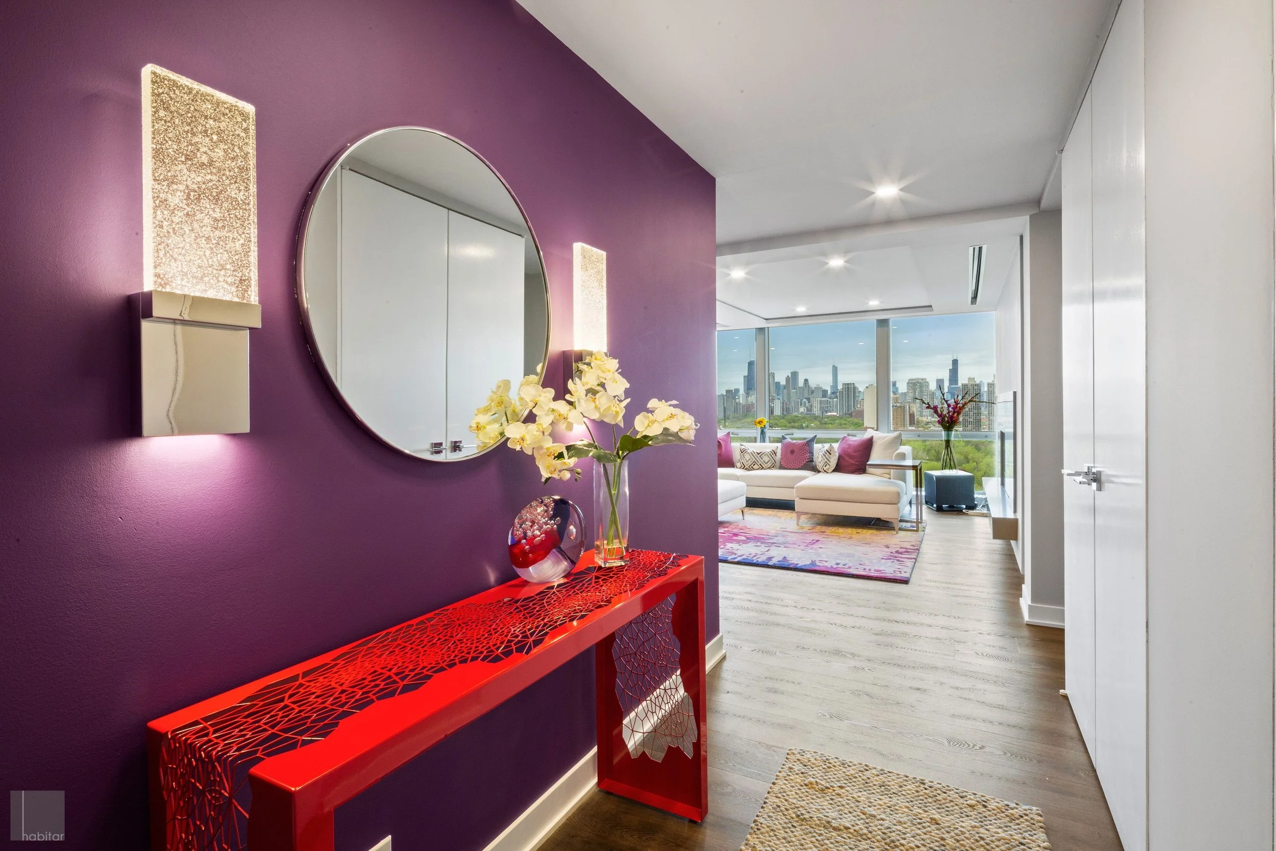 Modern living room with purple accent wall, decorative console table, white flowers, round mirror, and city view through large windows.
