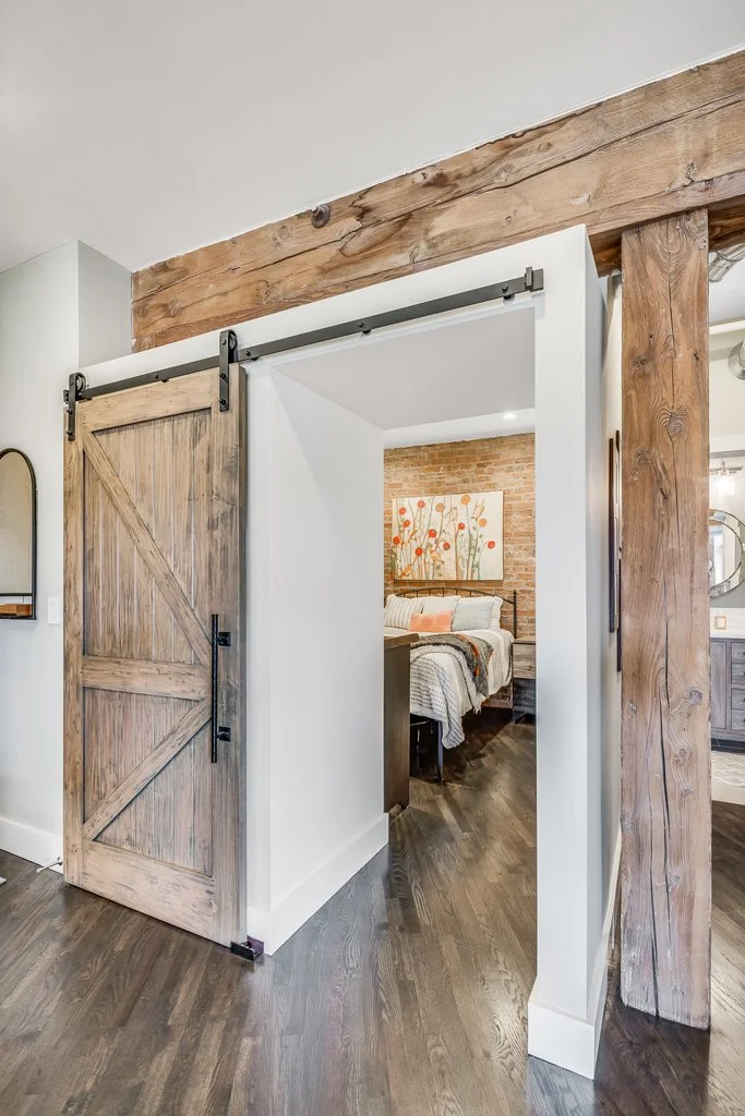 Interior view of a bedroom with a rustic sliding barn door made of weathered wood, mounted on black metal hardware, leading into a bedroom with an exposed brick wall, a bed with white and pastel bedding, and a floral painting on the wall.