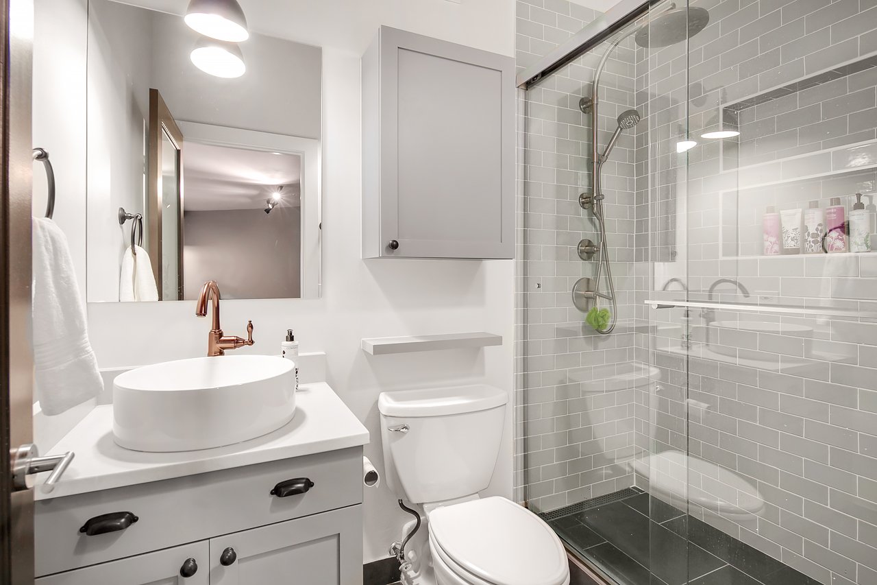 A modern bathroom with a white vanity, a vessel sink, a copper faucet, a mirror, a toilet, a wall cabinet, and a glass-enclosed shower with gray subway tiles and built-in shelves.