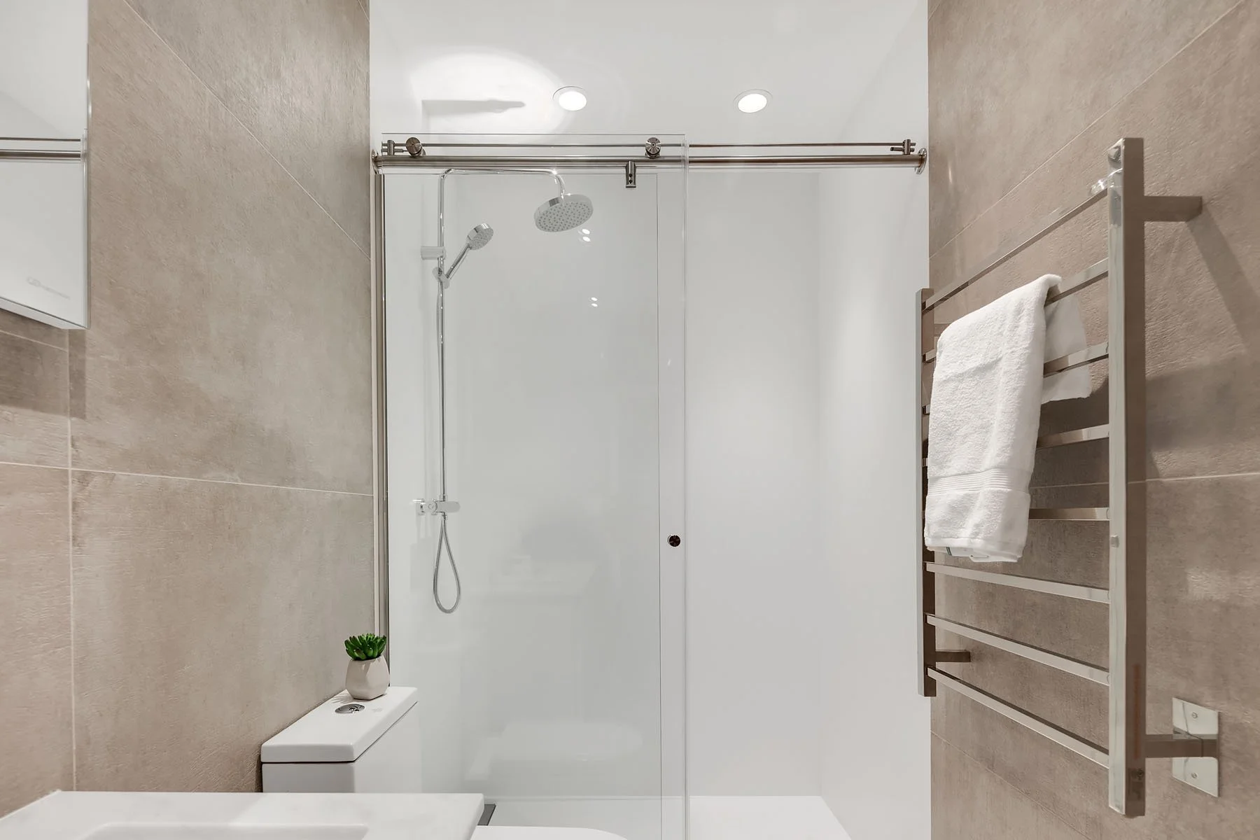 Modern bathroom with beige tiled walls and floor, glass shower enclosure with sliding door, rain showerhead, towel rack with white towel, small potted plant on toilet tank, white toilet and sink.