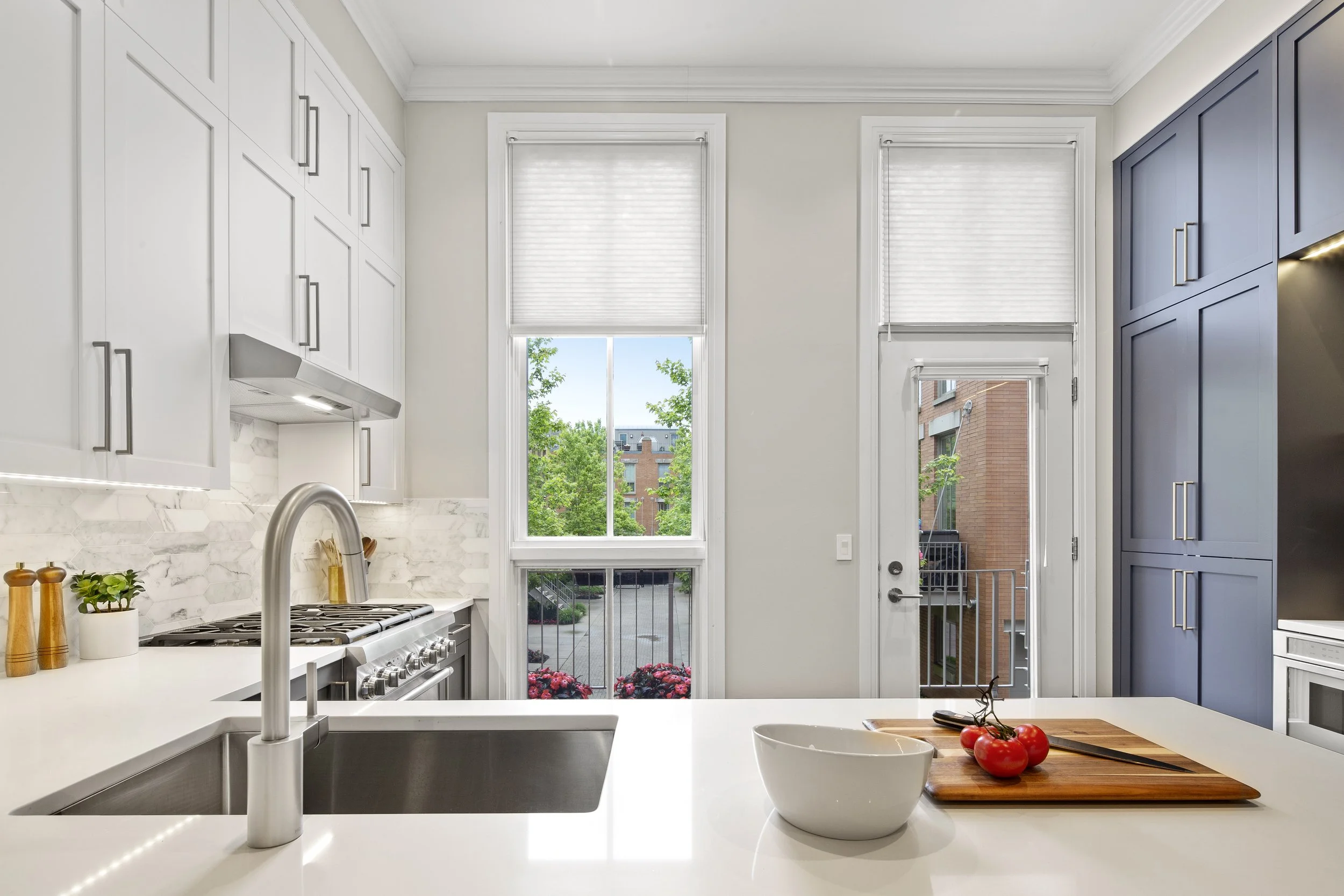 Modern kitchen with white and blue cabinets, large window, glass door, marble backsplash, kitchen sink, countertop with bowl and tomatoes, and outdoor view of trees and buildings.