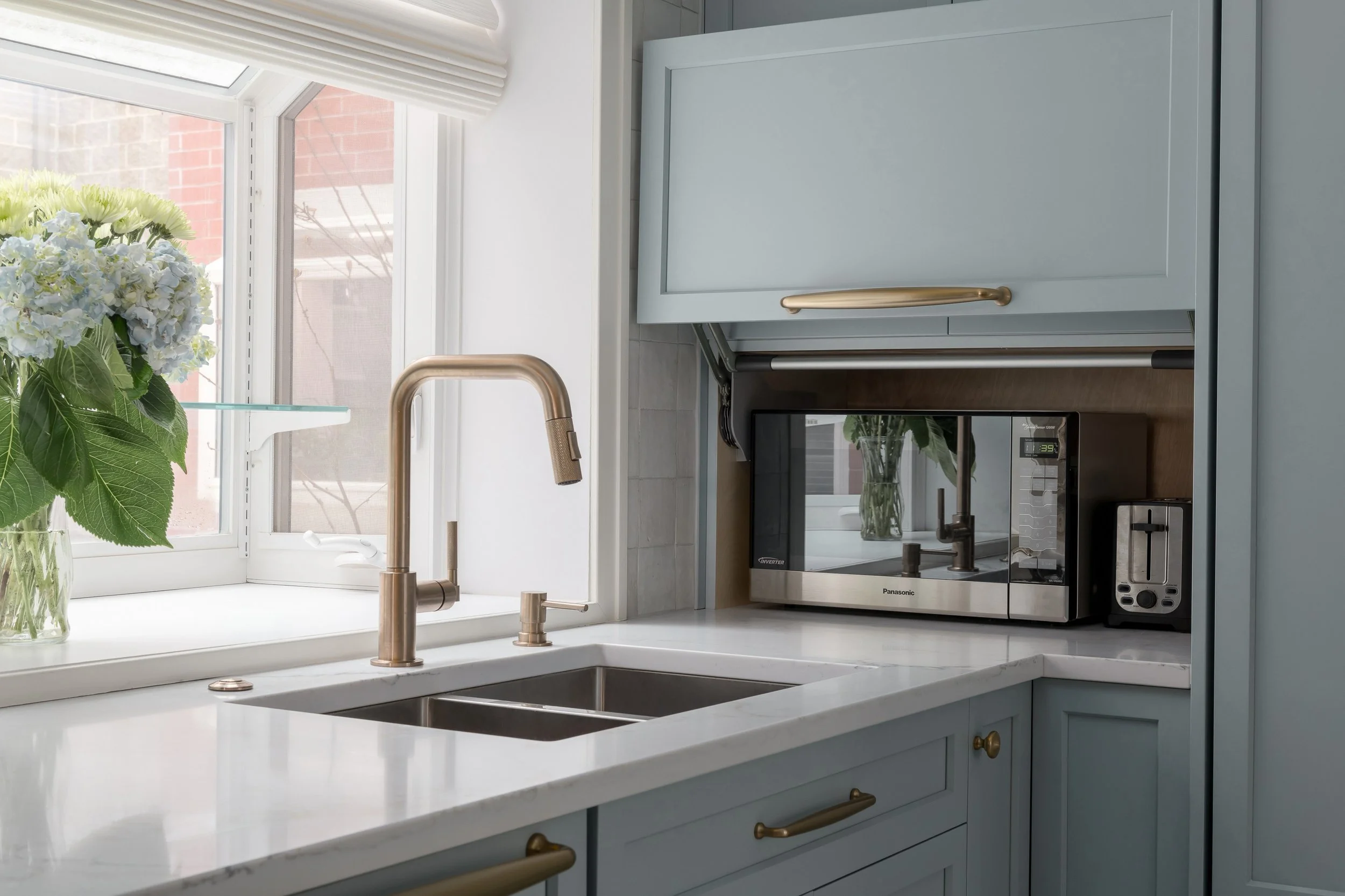 Kitchen with a light blue cabinet, a white marble countertop, a stainless steel faucet, a microwave oven, a toaster oven, and a window with white blinds.