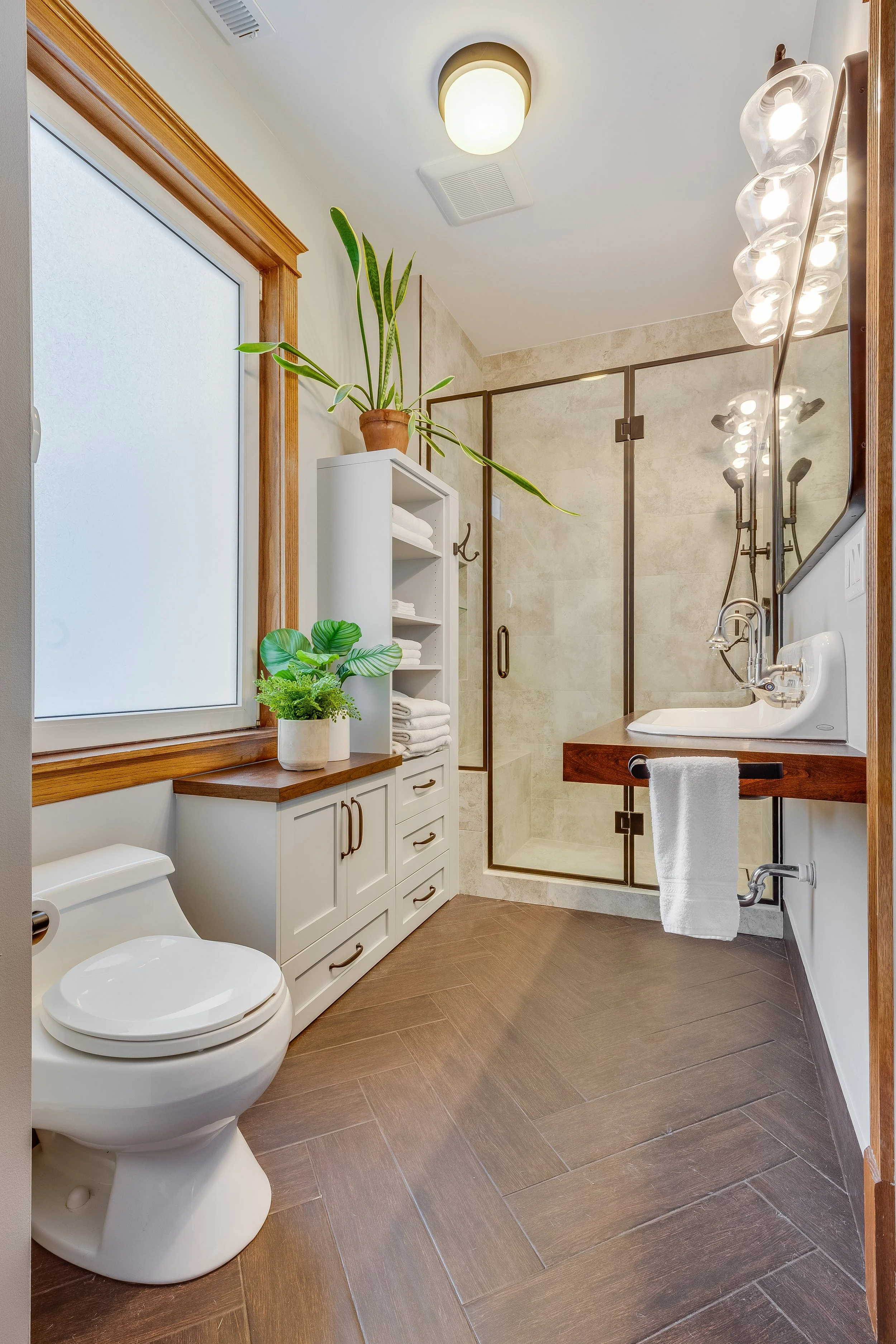 Modern bathroom with a freestanding cabinet, potted plants, a frosted window, a walk-in shower with glass doors, and a vanity with a wooden countertop and a mirror with multiple light bulbs.