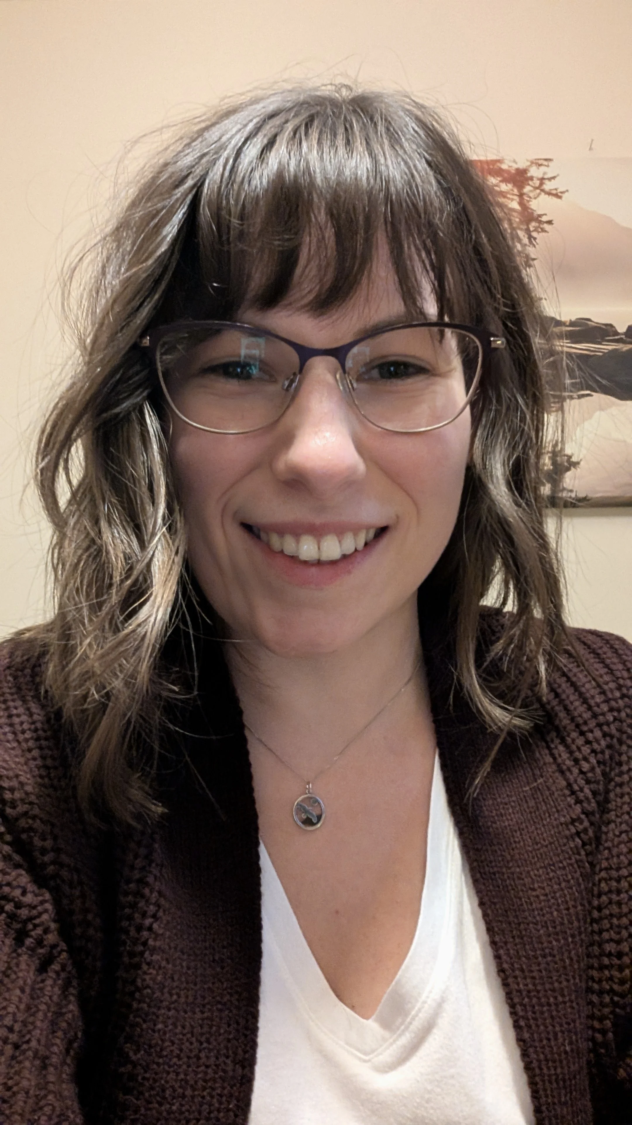 A woman with brown hair, glasses, and a necklace, smiling at the camera in an indoor setting.