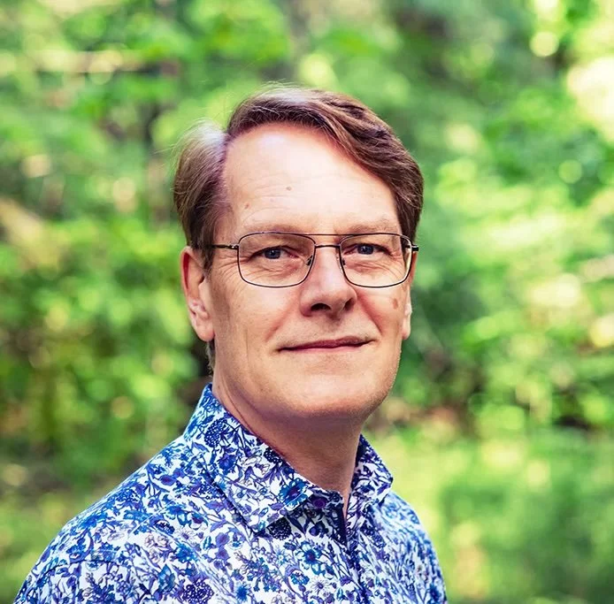 Portrait of a man with glasses wearing a blue patterned shirt outdoors surrounded by green trees.