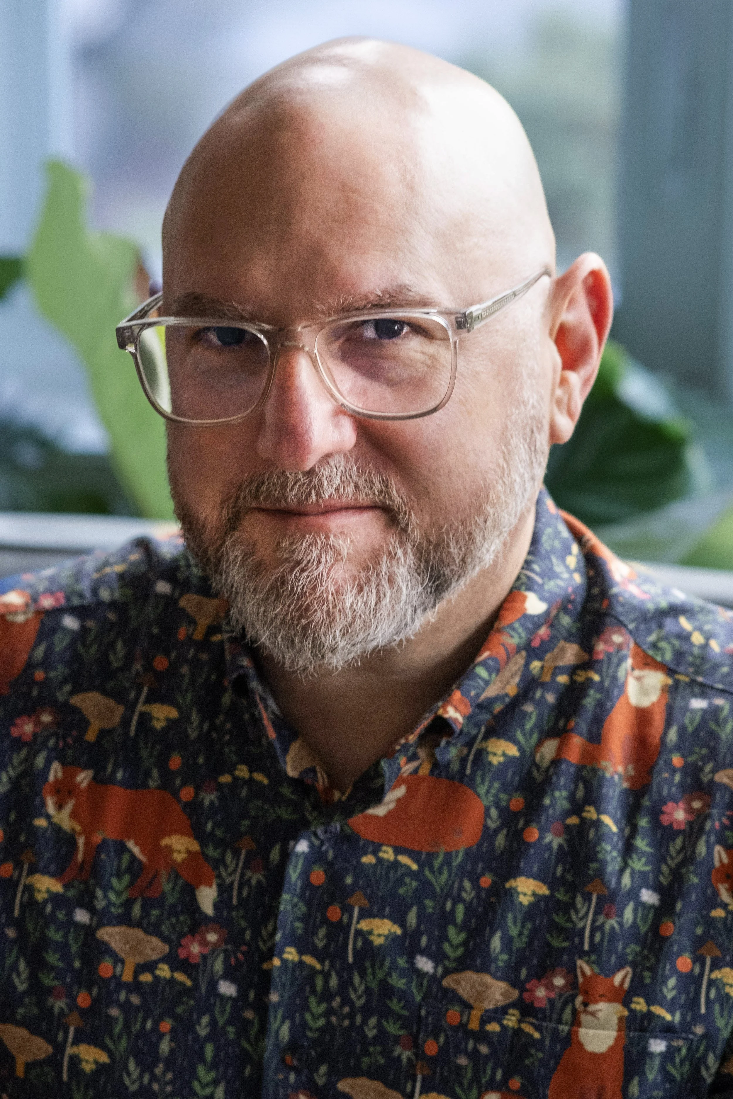 A middle-aged man with glasses, a beard, and a bald head, wearing a colorful shirt with fox and forest animal patterns, sitting indoors with a blurred green plant and window in the background.